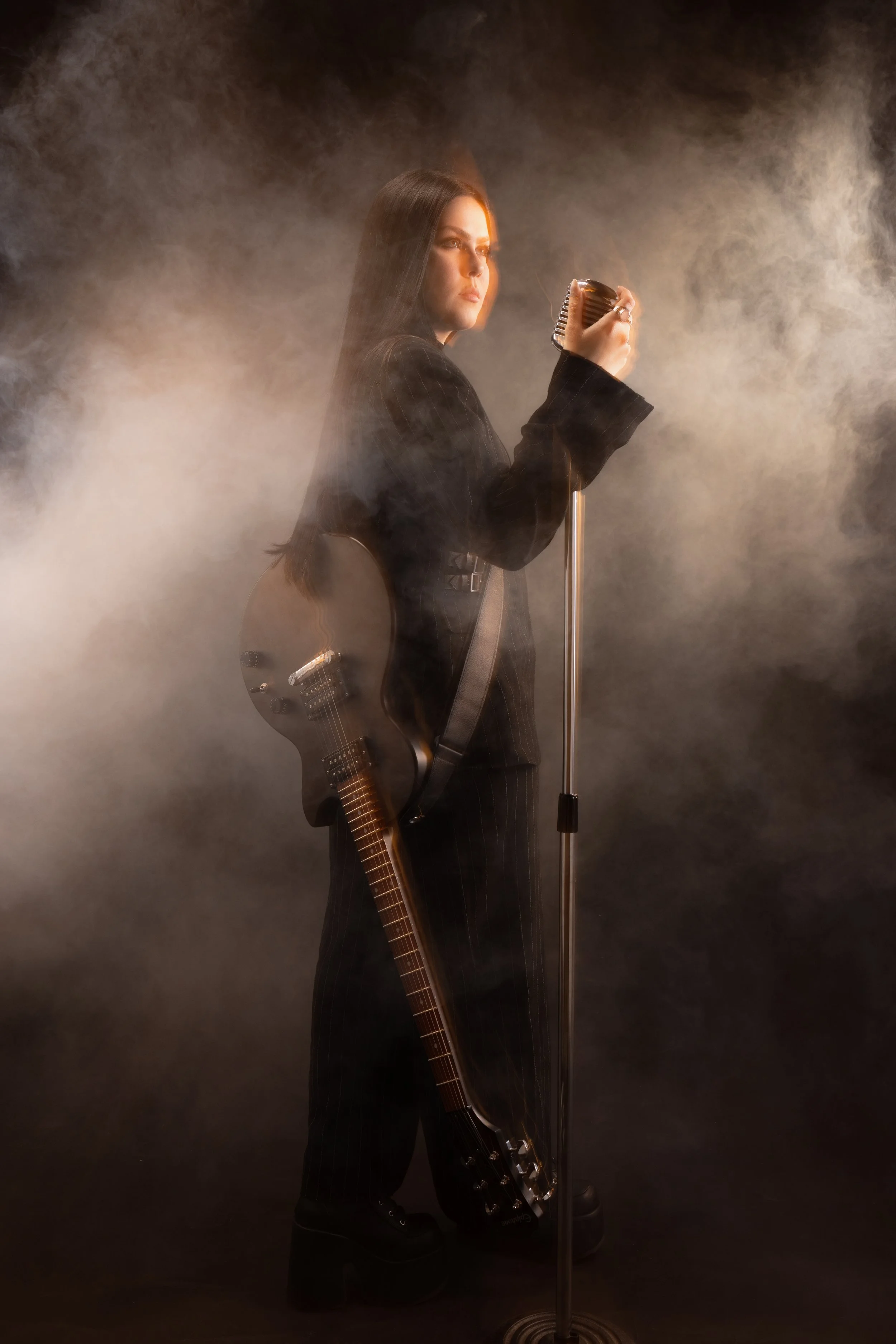 A woman with long dark hair wearing a black pinstripe suit, holding a vintage microphone, with a guitar strapped to her back, in a smoky atmosphere.