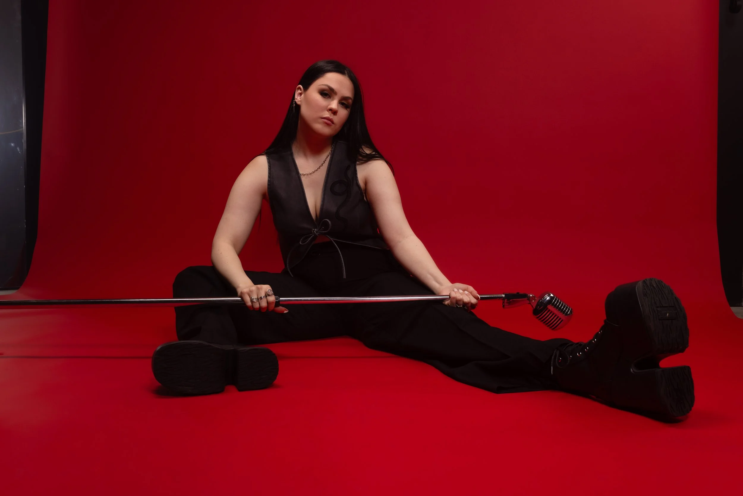 A woman with dark  hair, wearing black pants and a grey vest with a snake embroidered ,is sitting on the floor in front of a red backdrop. She holds a microphone and has a serious expression. There are black fabrics and a microphone stand around her.