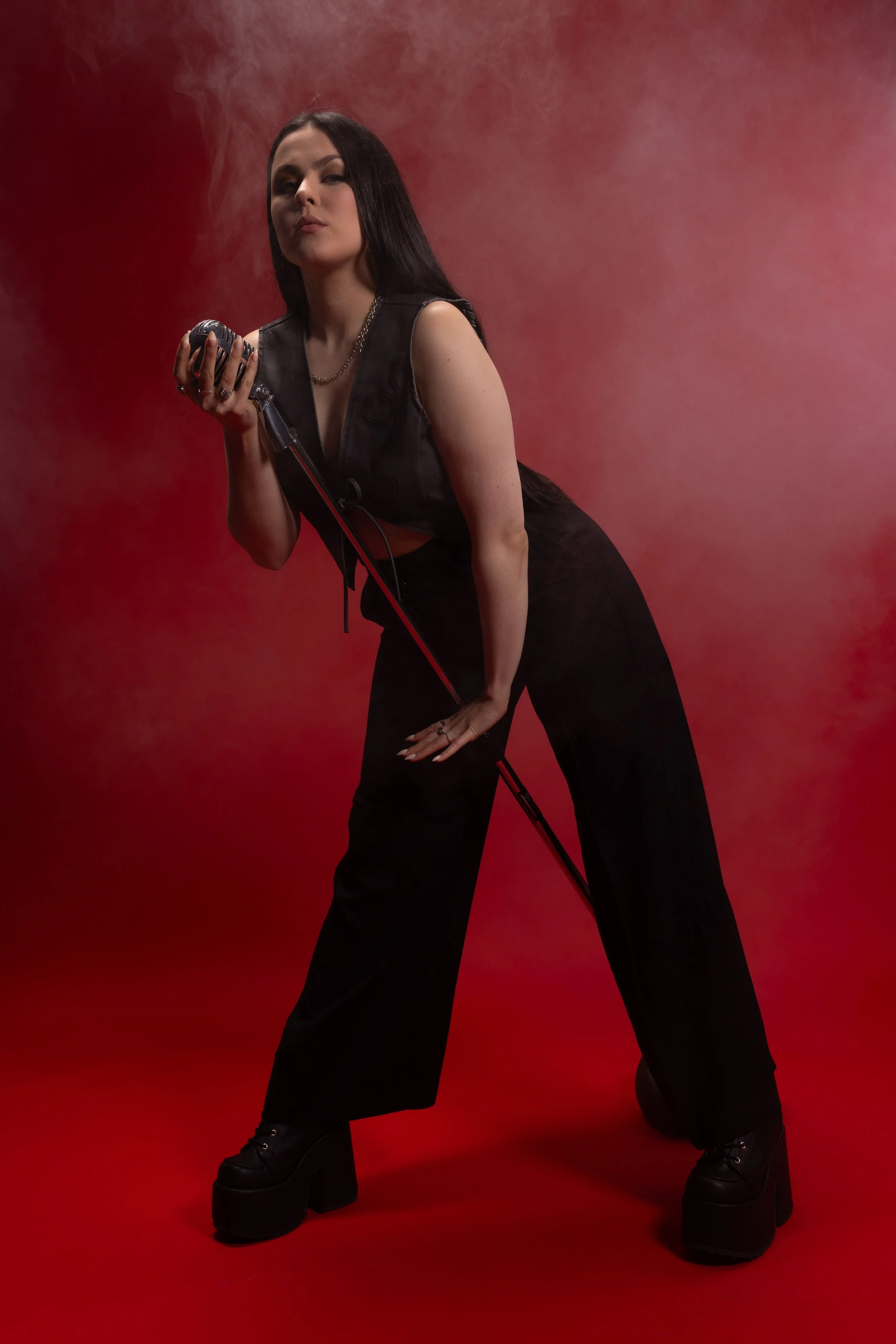 A woman with long dark hair wearing black platform shoes, black pants, and a grey vest with a snake embroider stands against a red background with smoke or mist in the air. She is holding a microphone in her right hand and has a confident expression.
