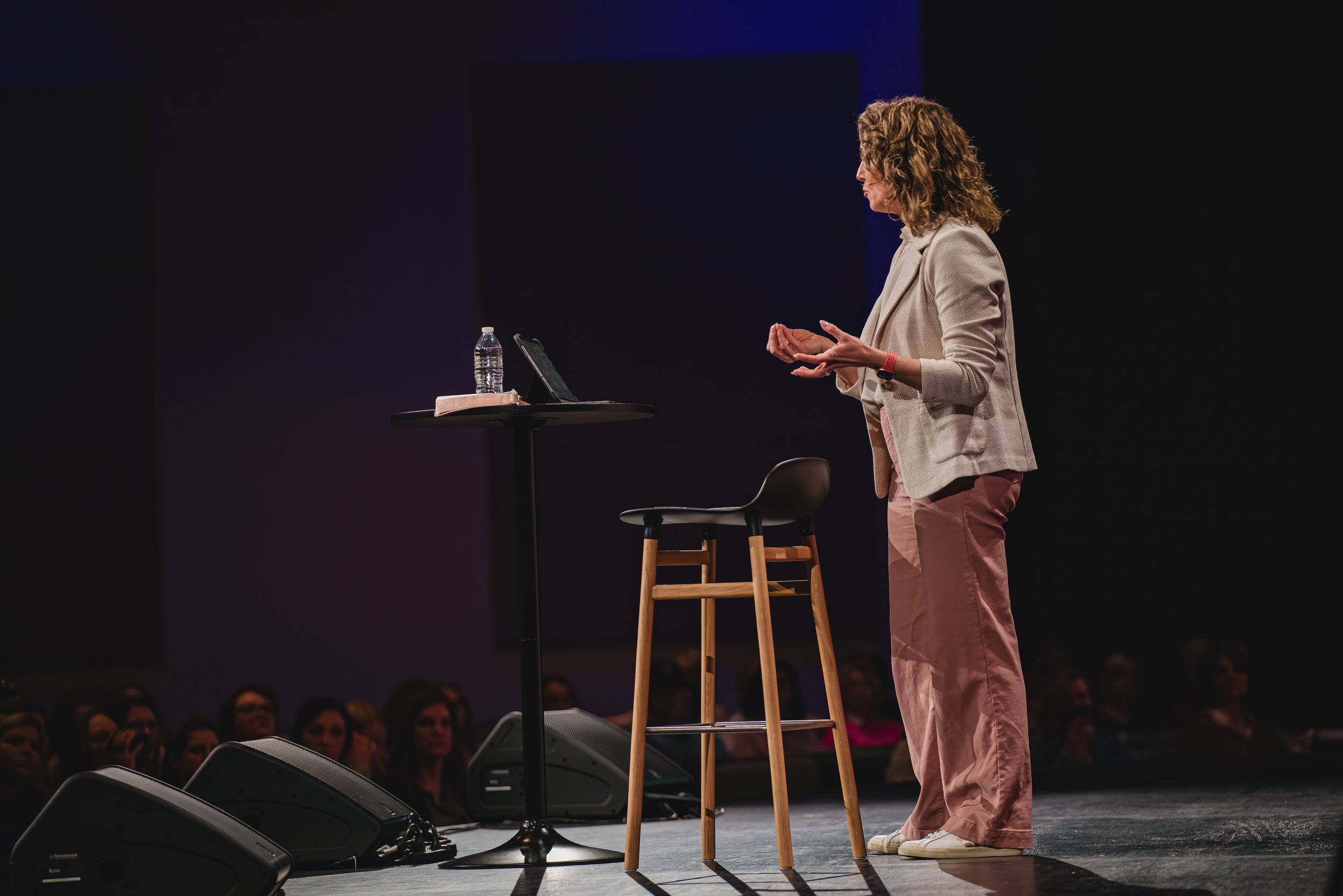 A woman with curly hair speaking on stage, standing next to a small round table with a water bottle, book, and tablet, with an audience in the background.