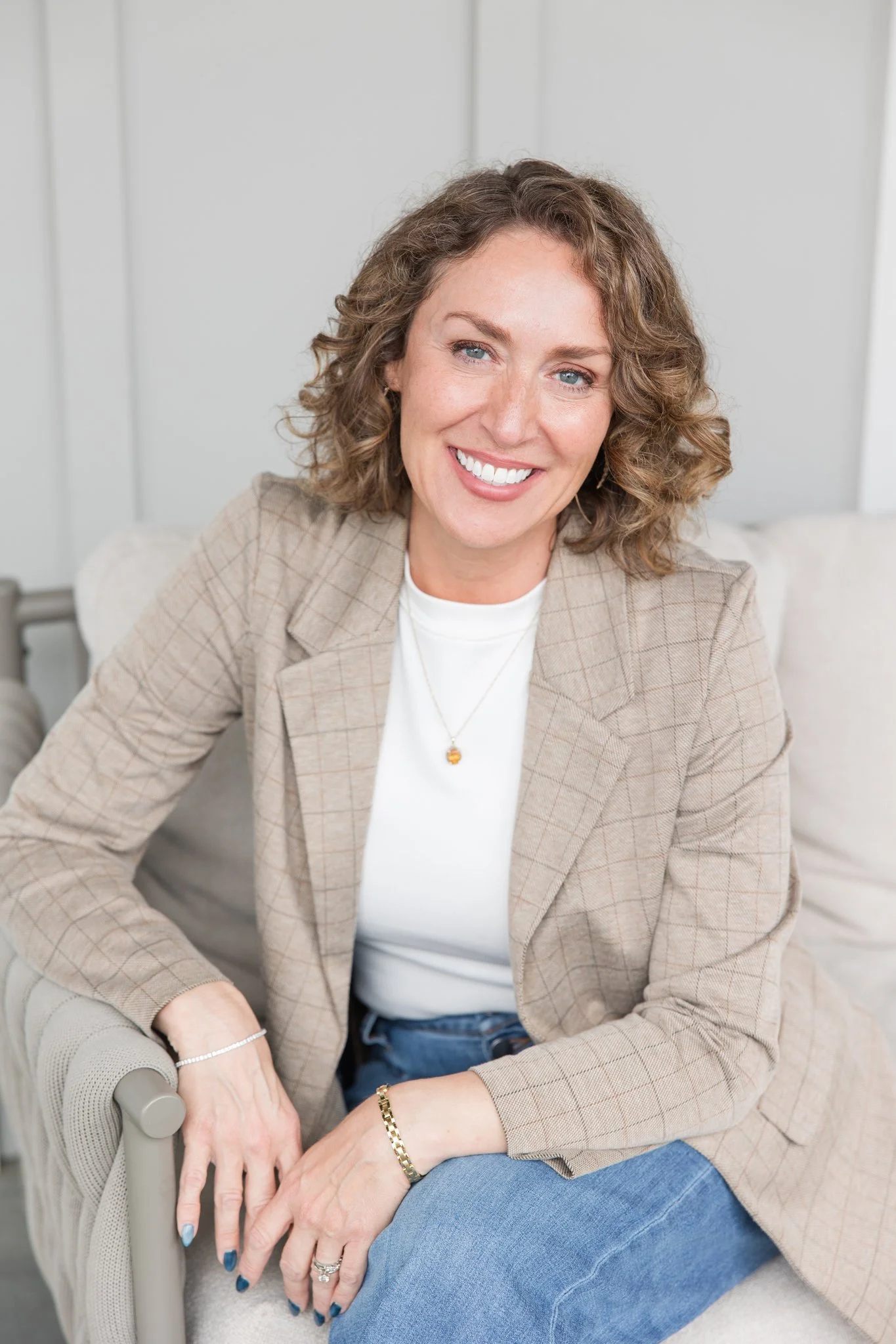 A smiling woman with curly light brown hair and blue eyes sitting on a beige couch, dressed in a beige plaid blazer, white top, and blue jeans, in a bright indoor setting.