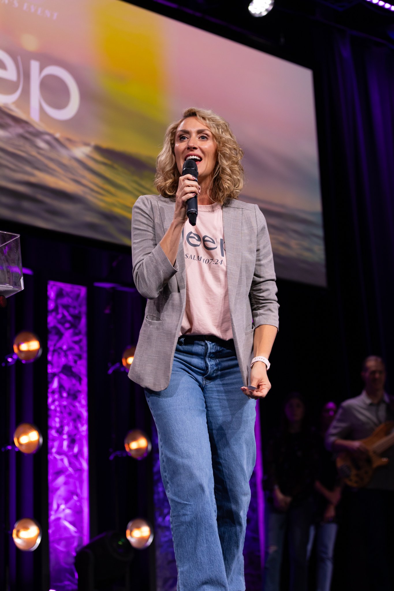 A woman with curly blonde hair, wearing a gray blazer, a light pink t-shirt, and jeans, is speaking into a microphone on stage. Behind her, there is a large screen displaying colorful abstract visuals. Purple lights illuminate the stage area.