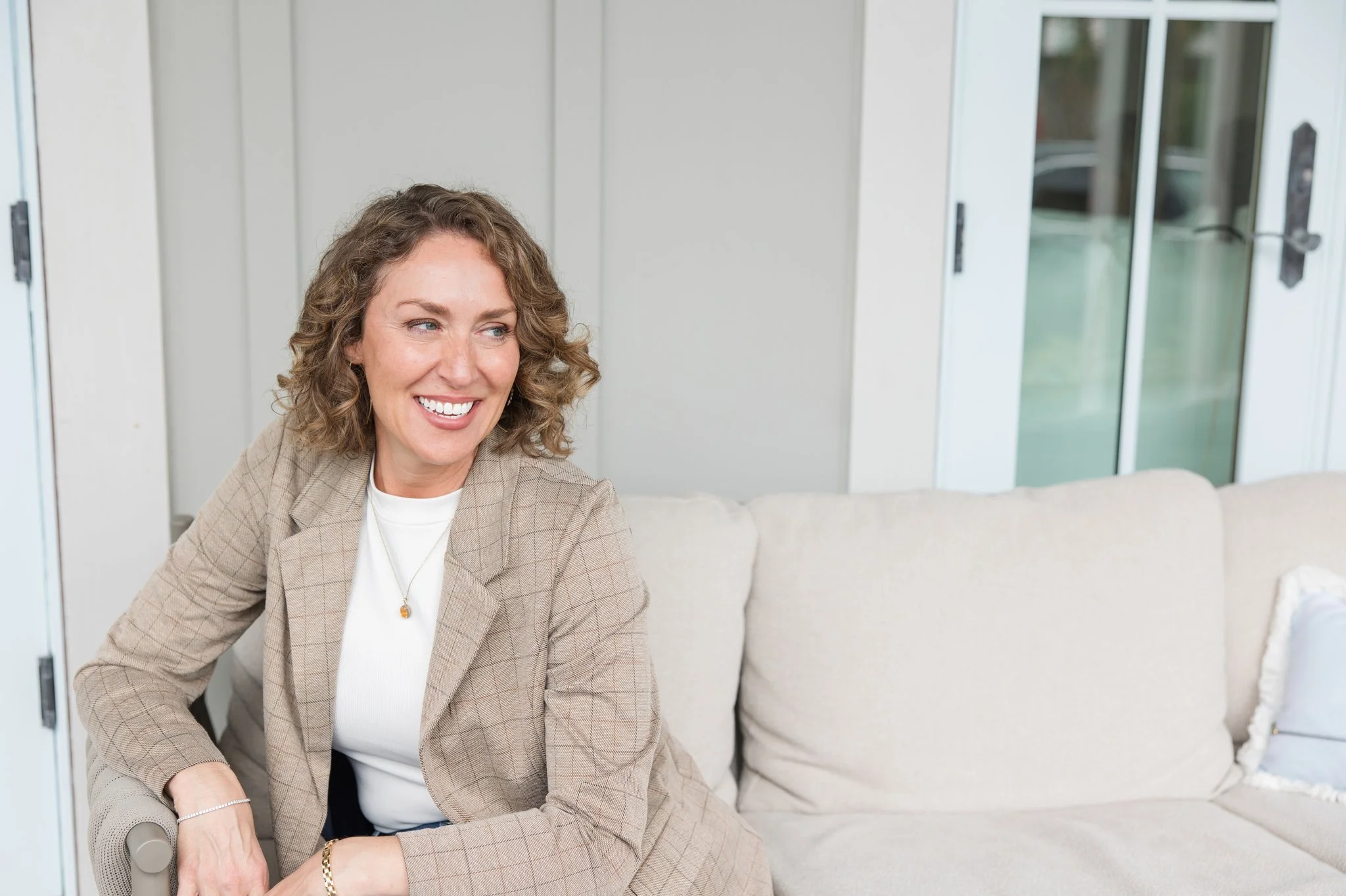A woman with curly brown hair smiling while sitting on a beige couch in a bright room with large glass doors.