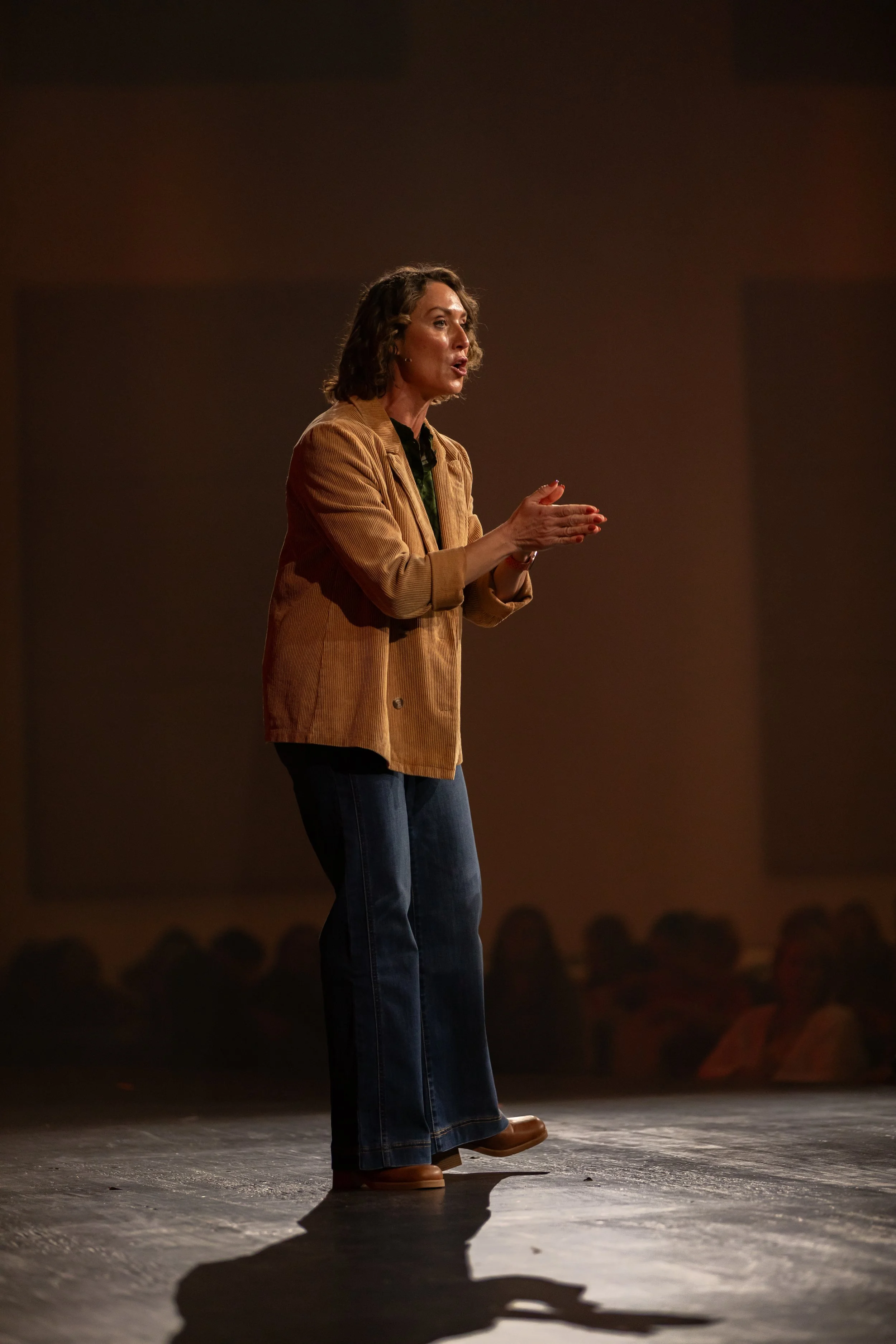 A woman giving a speech or performance on a stage, clapping her hands, wearing a tan blazer, black top, jeans, and brown boots.