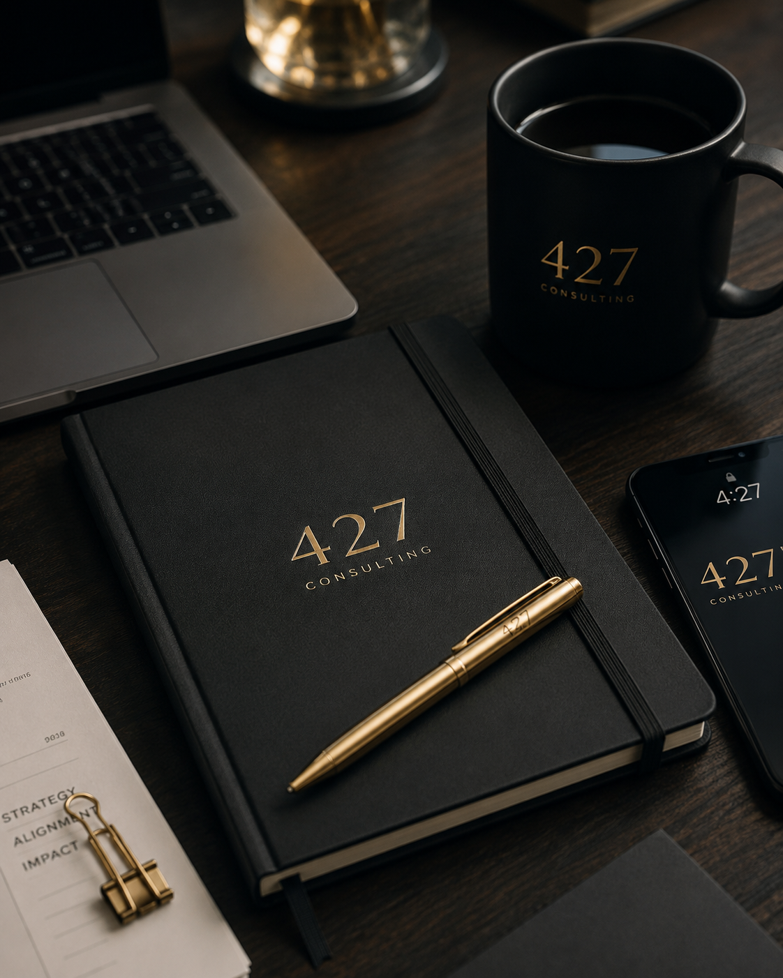 A black notebook, a gold pen, a coffee mug, a laptop, a smartphone, and a paper with a gold binder clip on a wooden desk. The notebook and mug both have the text '427 Consulting' in gold.