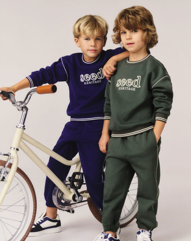 Two young boys wearing matching 'seed HERITAGE' sweatshirts and sweatpants, standing next to a bicycle, against a plain white background.