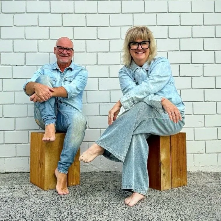 A smiling middle-aged man and woman sitting barefoot on wooden blocks against a white brick wall, both dressed in light blue denim outfits and glasses.
