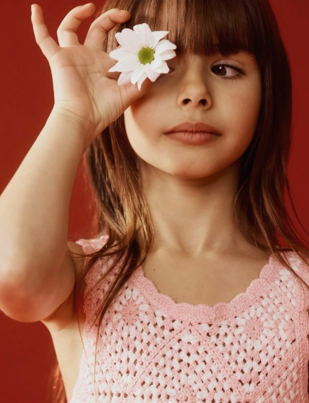 A young girl with red hair and a pink crocheted top holds a white flower with a green center over one eye against a red background.
