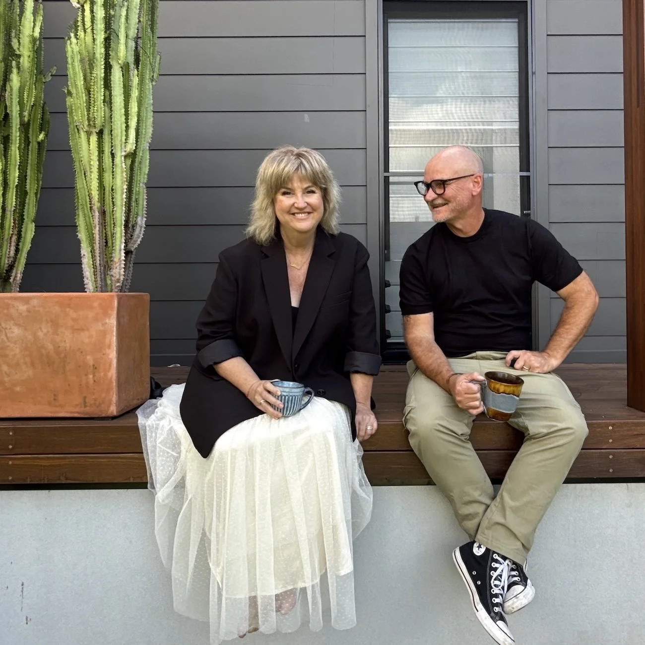 A woman and a man sitting on a wooden porch, each holding a tea cup. The woman is dressed in a black blazer and a white skirt, while the man is wearing a black t-shirt, beige pants, and black Converse sneakers. They are smiling and appear to be engaged in a conversation. There is a large potted cactus plant on the left side of the image and a gray house exterior in the background.