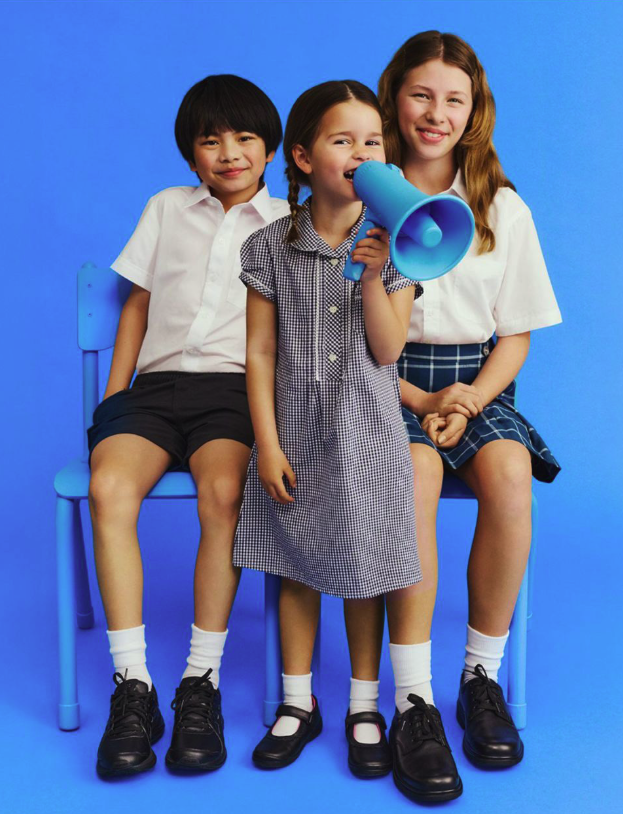 Three children sitting on a blue bench against a bright blue background. The girl in the middle holds a blue megaphone, smiling, as the other two children sit on either side of her. The children are dressed in school uniforms.