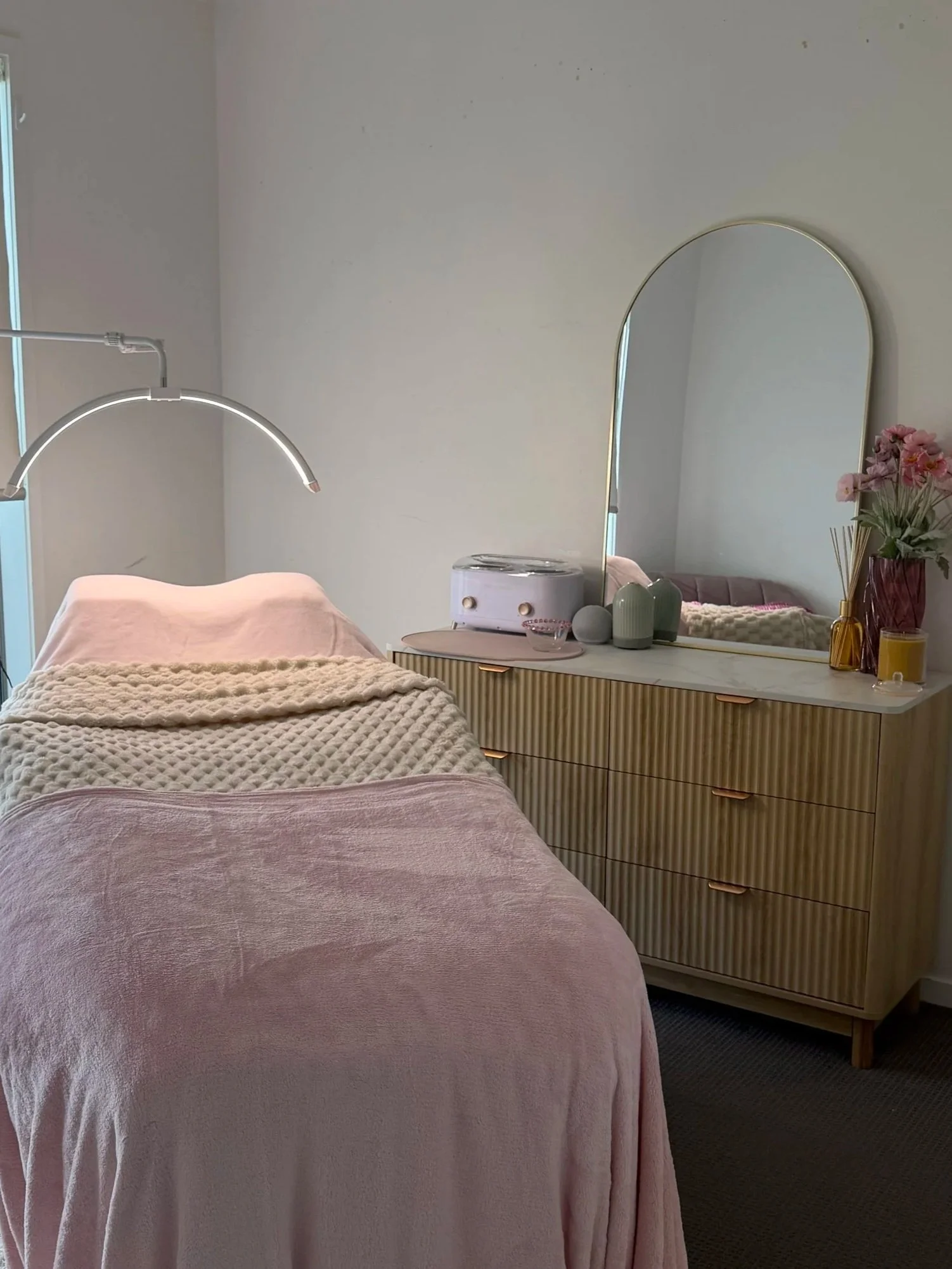 Pink blanket on a bed in a bedroom with a wooden dresser, a mirror, and decorative items, including a flower vase with pink flowers, on top of the dresser.