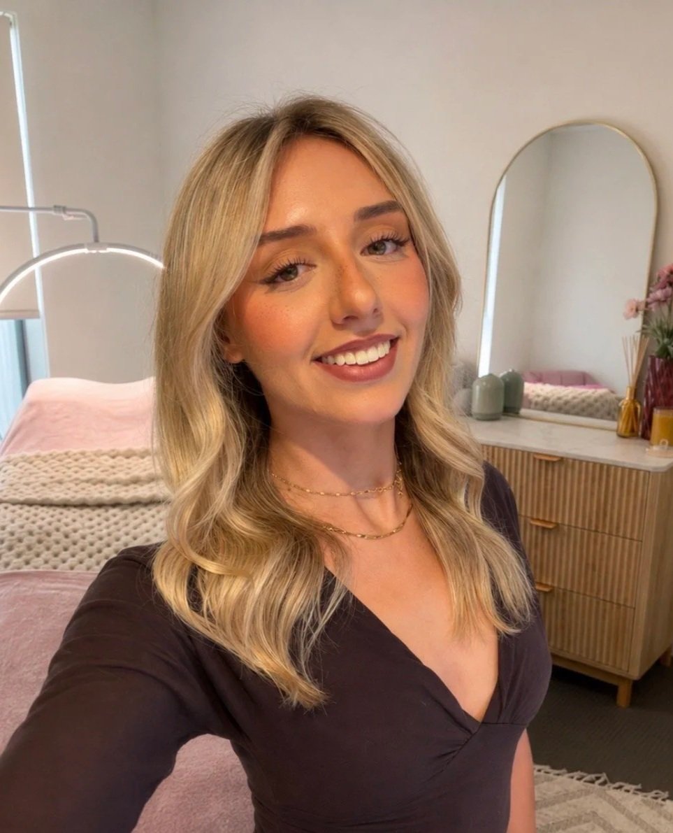 A woman with blonde hair styled in loose waves taking a selfie in a cozy, modern bedroom. She is smiling and wearing a black top with layered gold necklaces. The background features a mirror, a dresser, and decorative items including plants and candles.