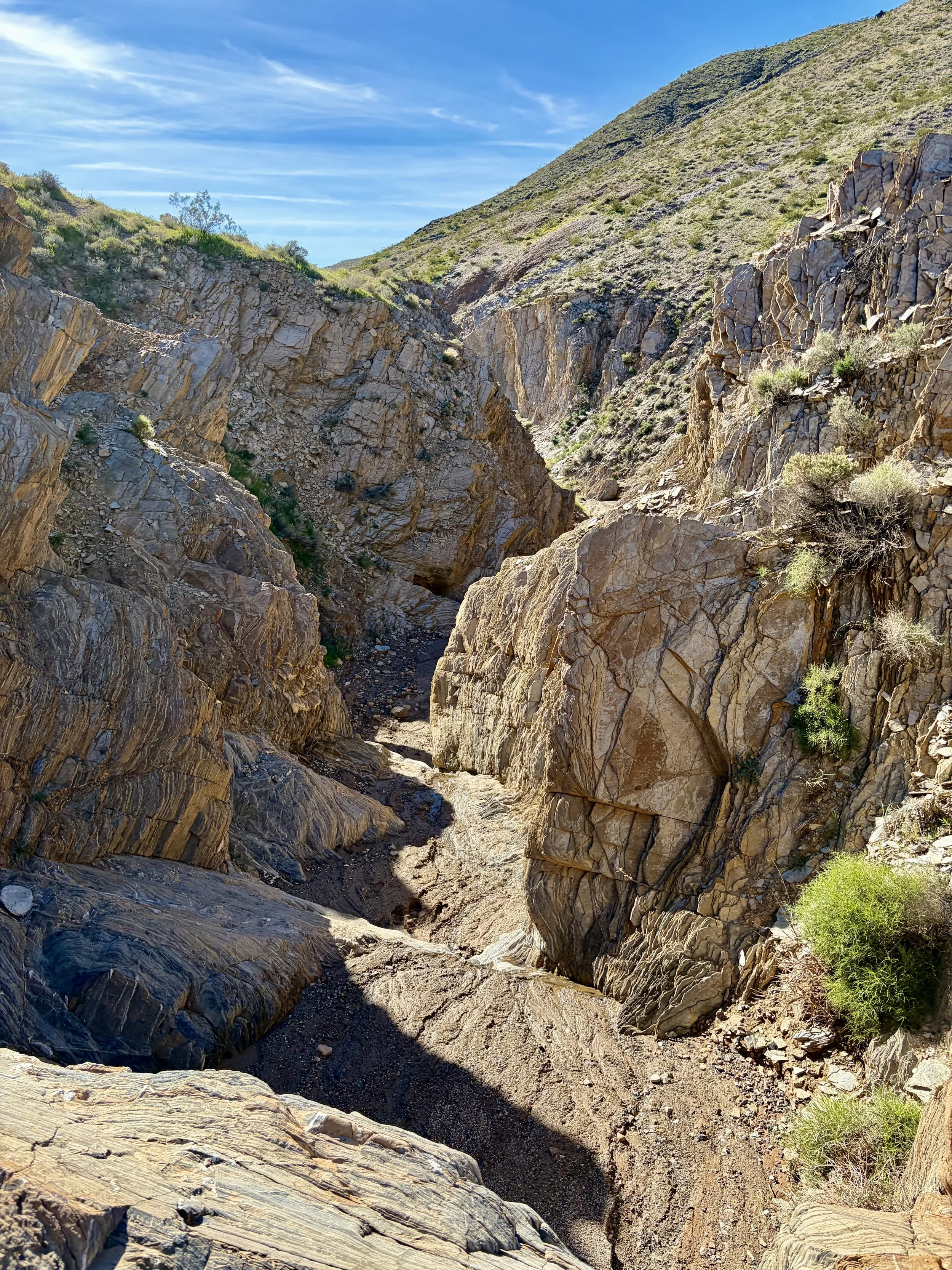 A dry, rocky canyon with steep, rugged stone walls and sparse green vegetation under a blue sky with wispy clouds.