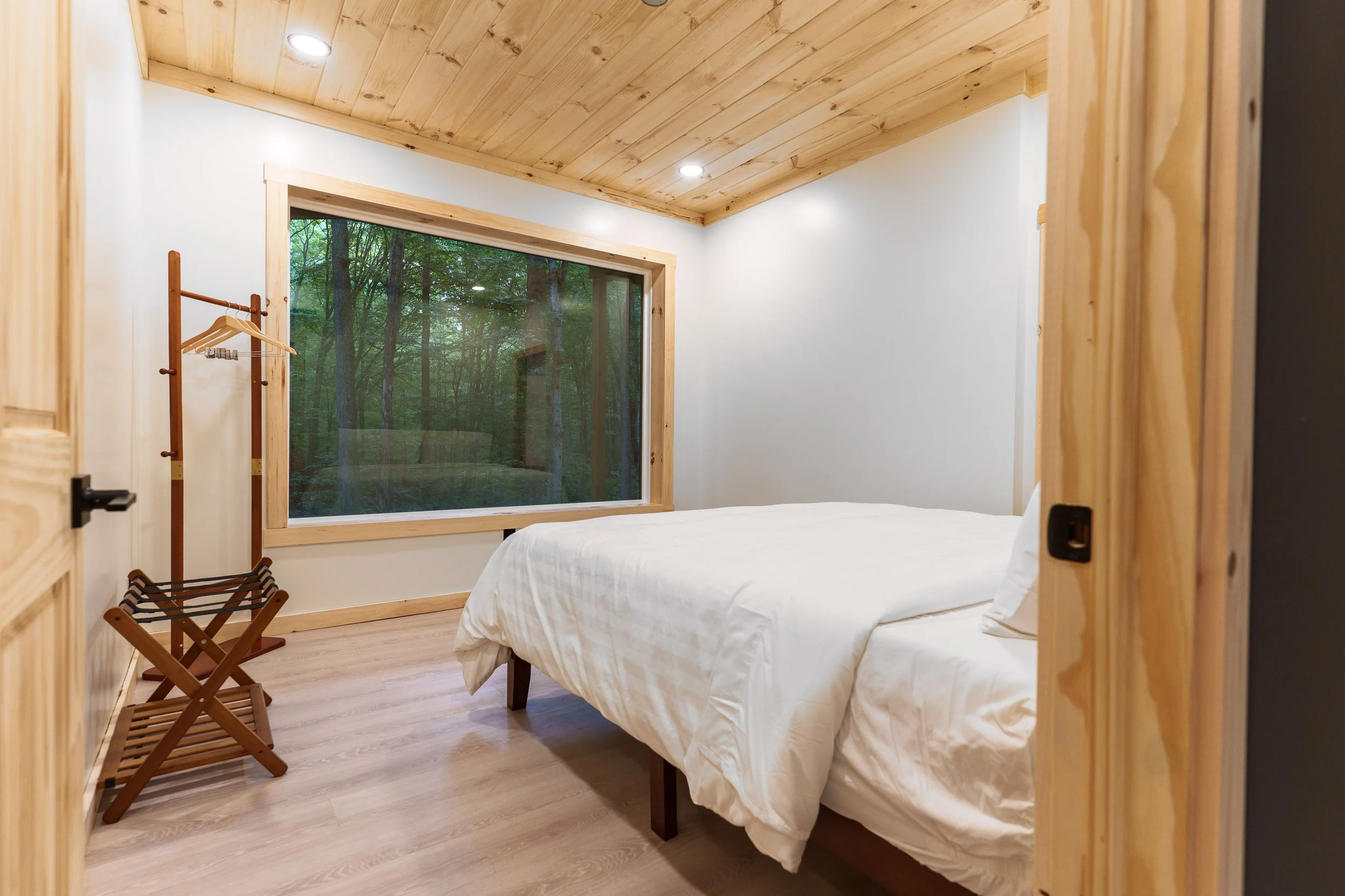 A cozy bedroom with a large window overlooking a lush forest. The room features wooden ceiling and trim, a white bed, a small wooden luggage rack, and a wooden clothes stand with hangers.