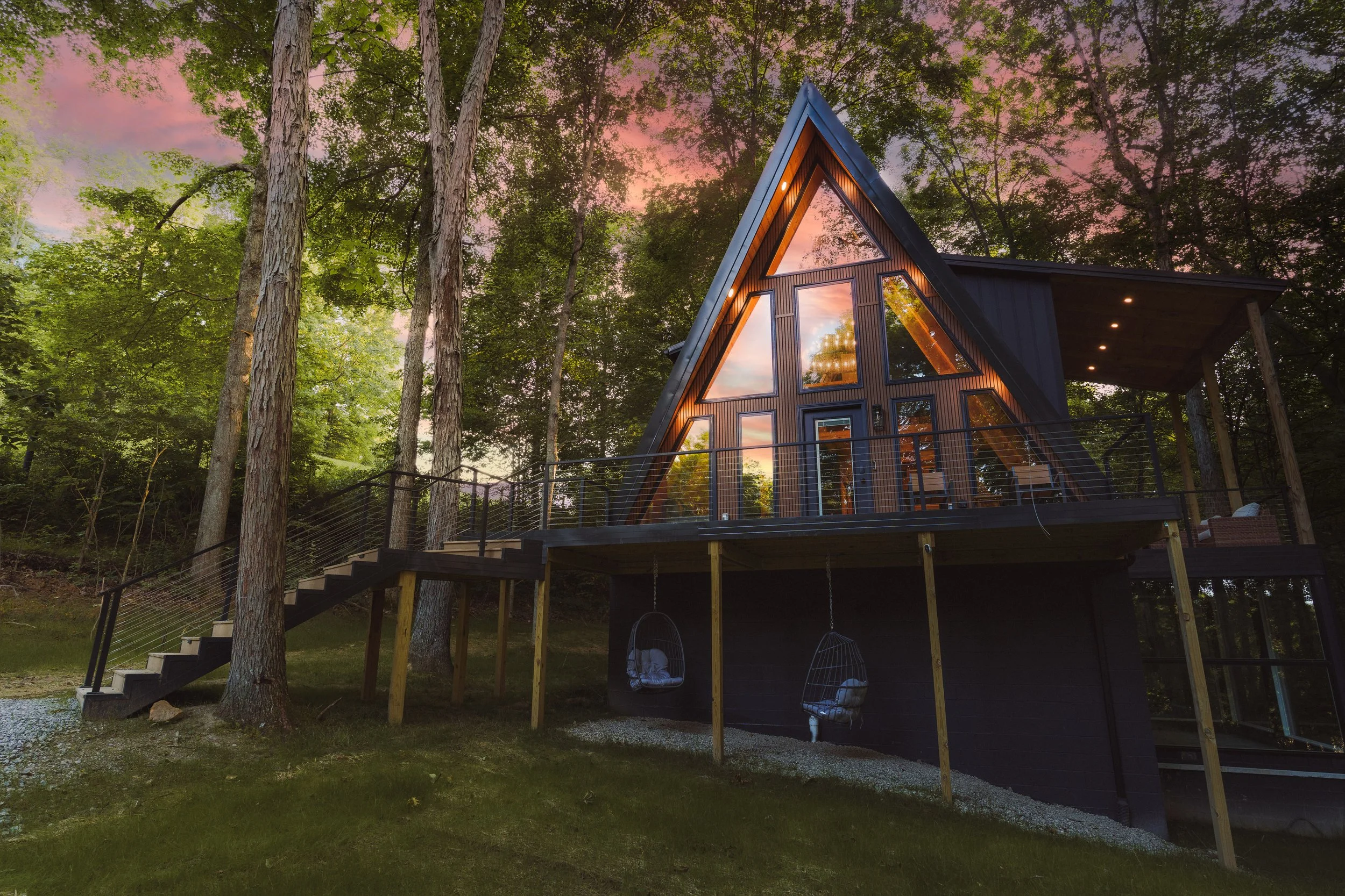 A modern A-frame house surrounded by tall trees and a sunset sky, with an elevated deck and hanging chairs underneath.