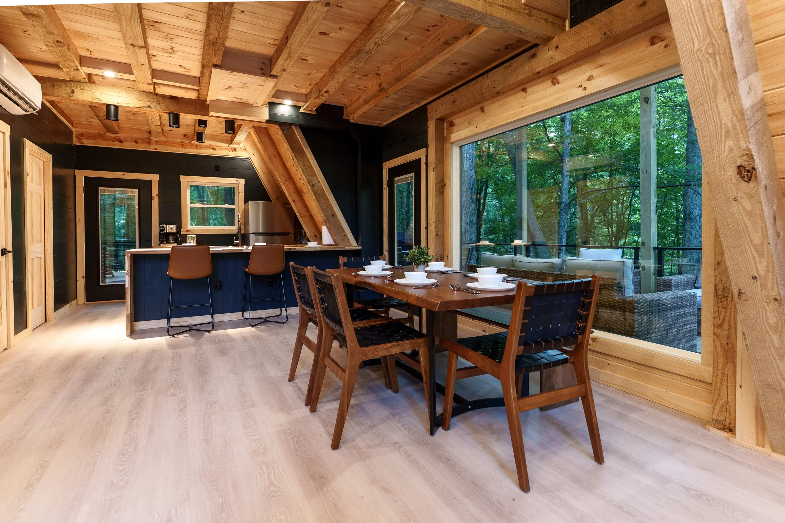 Interior of a wooden house with large windows showing trees outside. It features a dining table with six chairs and a kitchen area with black walls, a counter, a fridge, and kitchen appliances. The ceiling and walls are made of natural wood.