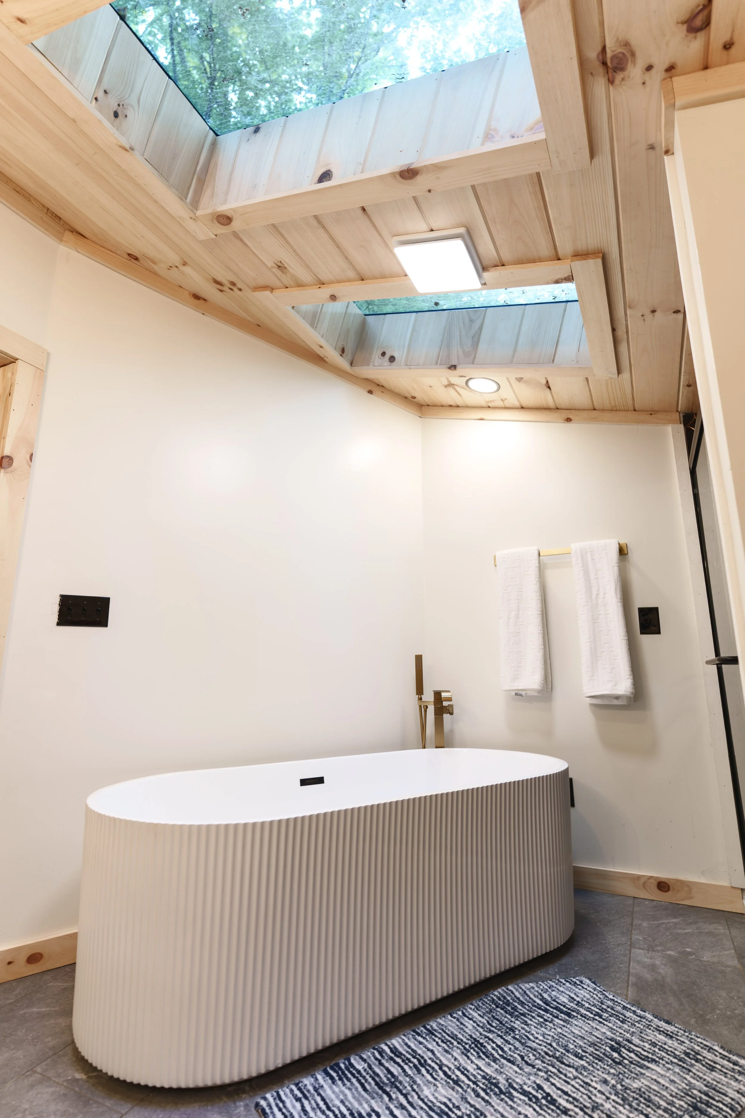Modern bathroom with a freestanding oval bathtub that has a ribbed exterior and a gold faucet. The ceiling features a wooden panel with two skylights and a square ceiling light. Towels are hanging on a gold towel bar, and a gray and white rug lies on a tiled floor.