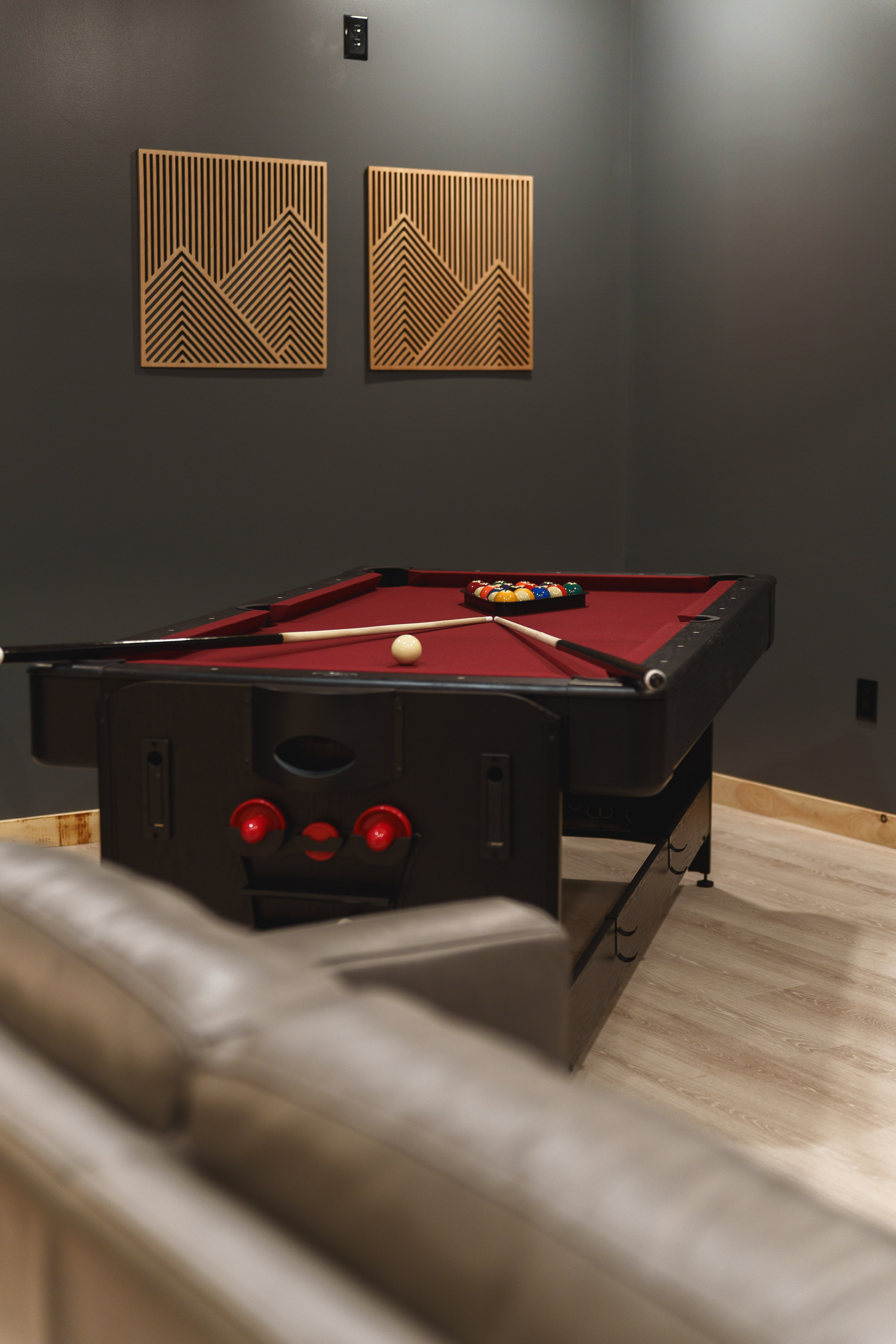 A pool table with billiard balls racked at one end and cue sticks resting along the sides, set in a room with dark gray walls and modern wooden wall art.