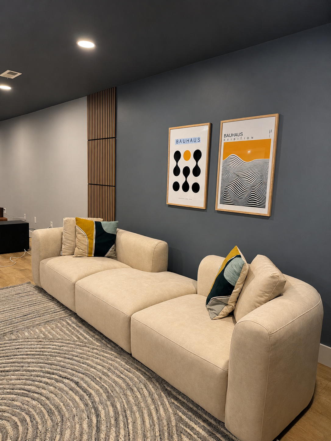 Modern living room with beige sofa, decorative pillows, dark blue wall, framed Bauhaus poster art, wooden accents, and a patterned rug.