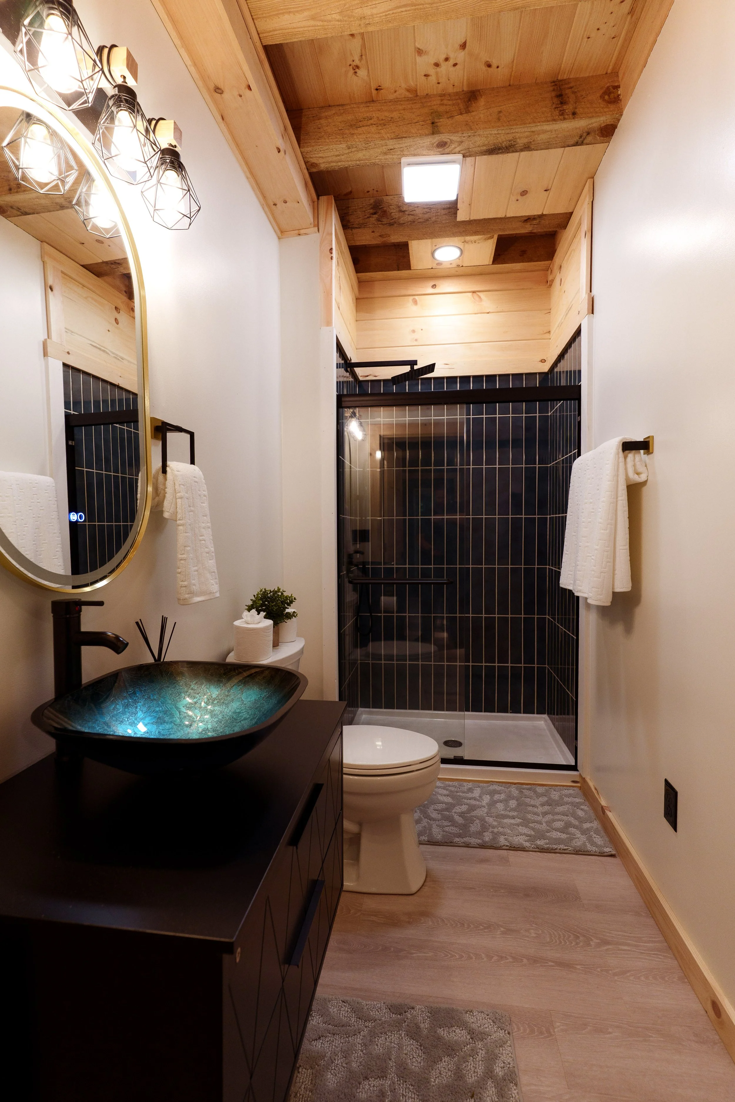 A modern bathroom with a black vanity, a unique blue-green vessel sink, a round mirror, a toilet, a small plant, a white towel hanging on a black towel bar, a glass-enclosed shower with dark tiles and a wooden ceiling overhead, and a patterned rug on the wooden floor.