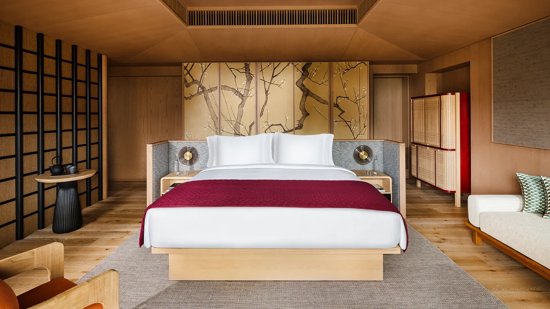 A modern bedroom with a bed that has white pillows and a burgundy quilt. Behind the bed is a gold and black artistic mural of tree branches. On each side of the bed are small nightstands with lamps and glasses of water. To the left, a small round black side table holds a teapot and cups. On the right side, a tall wooden cabinet with woven front panels is visible. The room has wood-paneled walls and flooring, with contemporary furniture and decor.