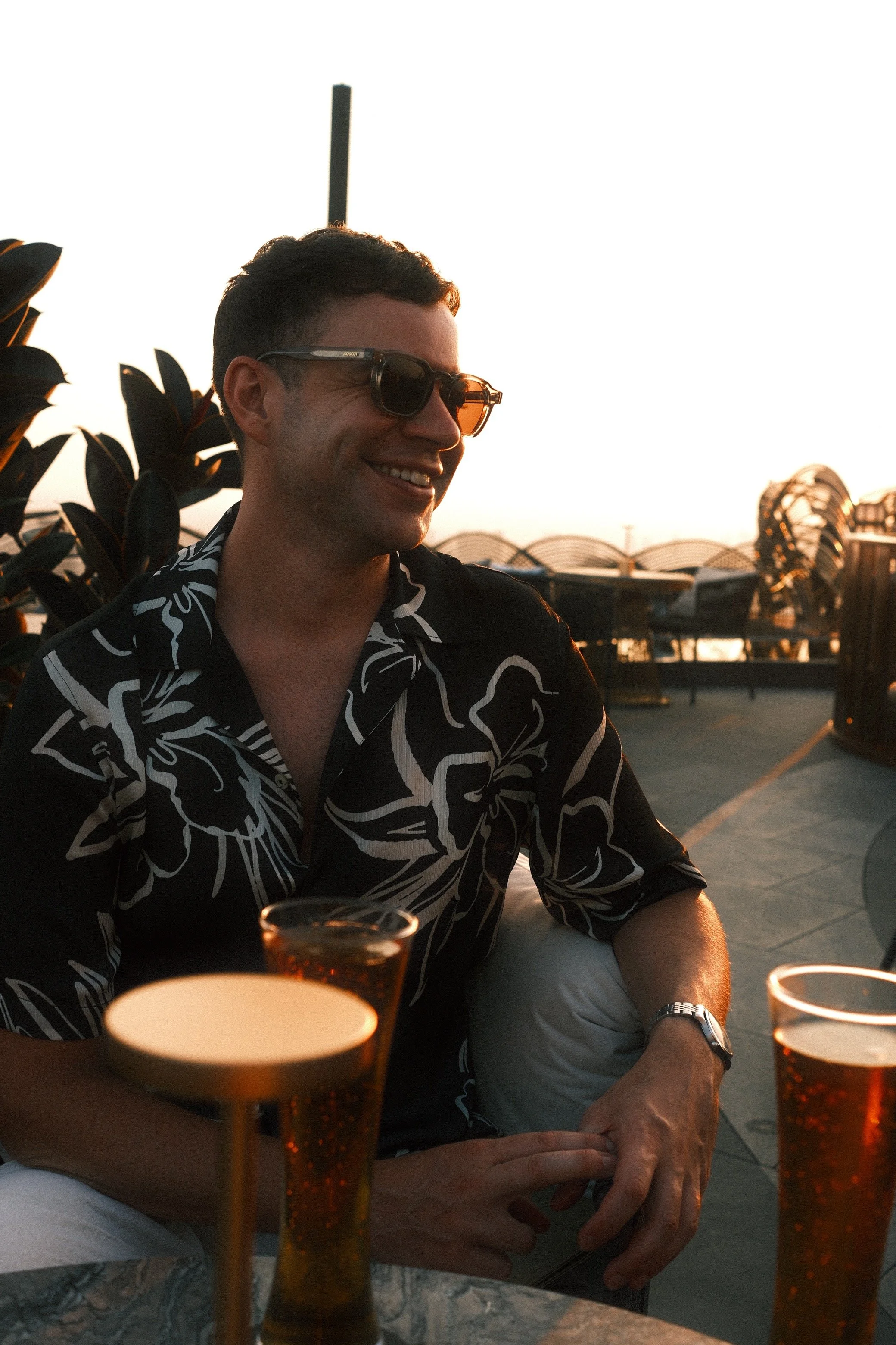 A man wearing sunglasses and a black shirt with a white floral pattern smiling while sitting at an outdoor table during sunset, with a drink in front of him and a modern structure in the background.