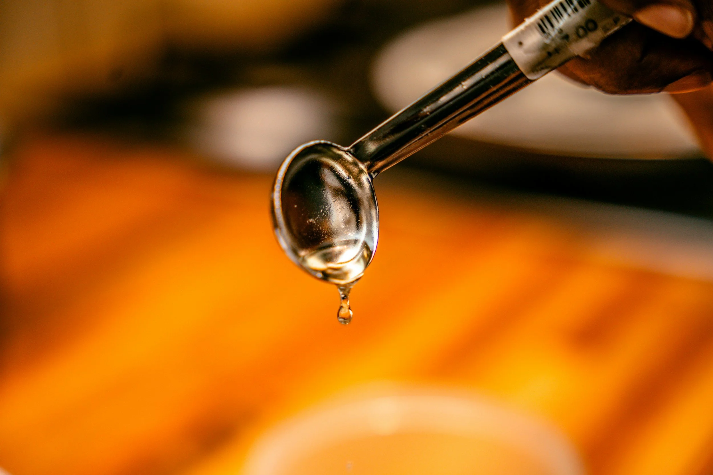 Close-up of a drop of clear liquid hanging from the tip of a metal needle, with blurred warm orange and brown background.