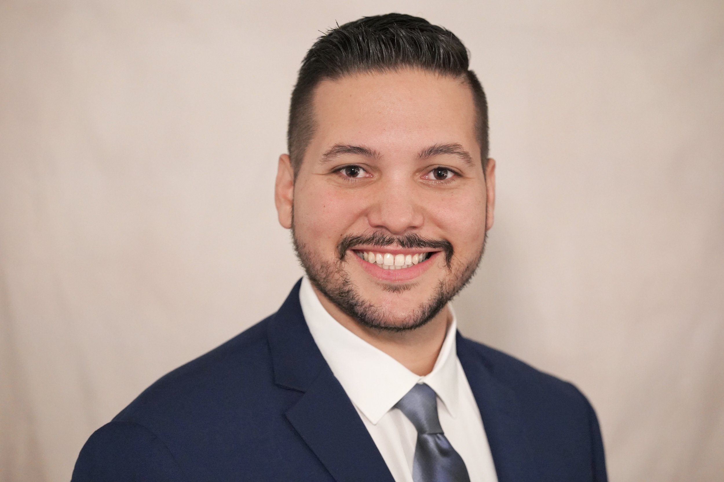 Headshot of a smiling young man with dark hair, a beard, and a mustache, wearing a navy blue suit, white shirt, and a gray tie, against a plain light-colored background.