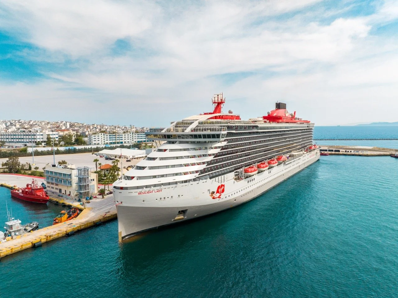 Large white cruise ship named Resilient Lady docked at a harbor with a small red ship nearby. The harbor is adjacent to a city with white buildings, and the water is calm under a partly cloudy sky.