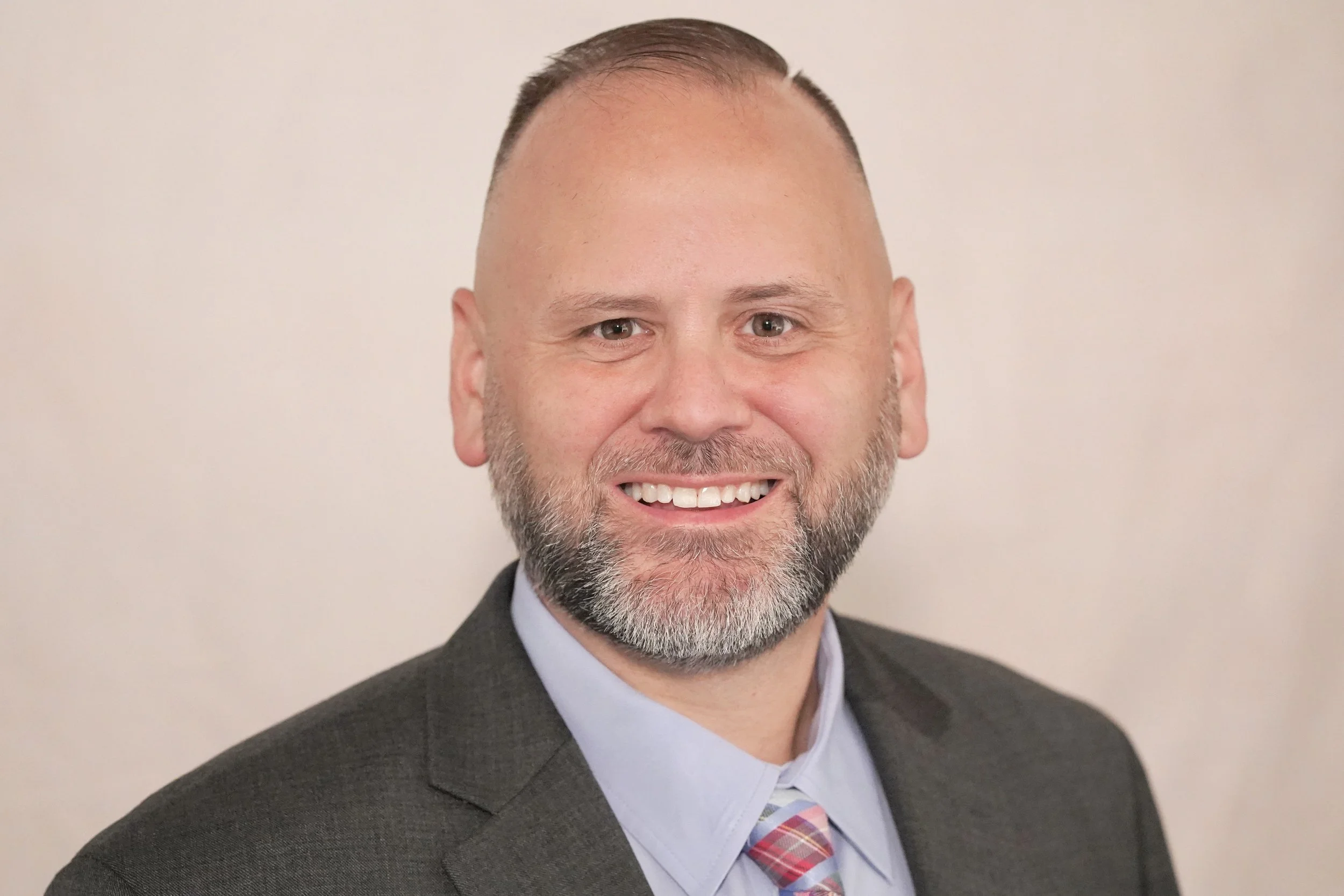 A middle-aged man with a beard and gray hair smiling, dressed in a gray suit, light blue shirt, and striped tie, against a plain light-colored background.