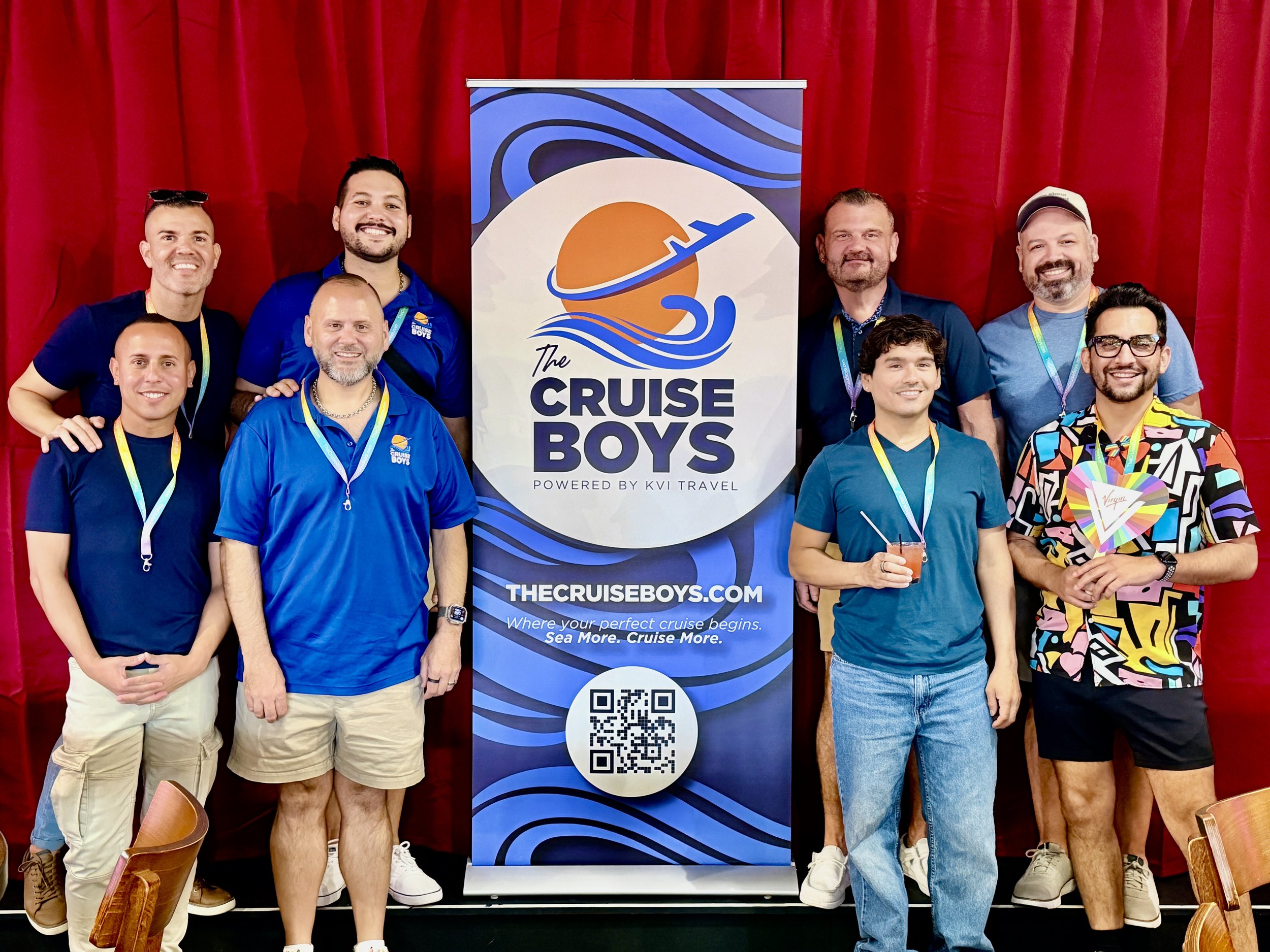 A group of nine men standing in front of a red curtain, posing for a photo. In the center, a vertical banner reads 'The Cruise Boys' with a logo of a sun, waves, and an airplane, and a QR code at the bottom. The men are smiling, dressed casually, and wearing lanyards.