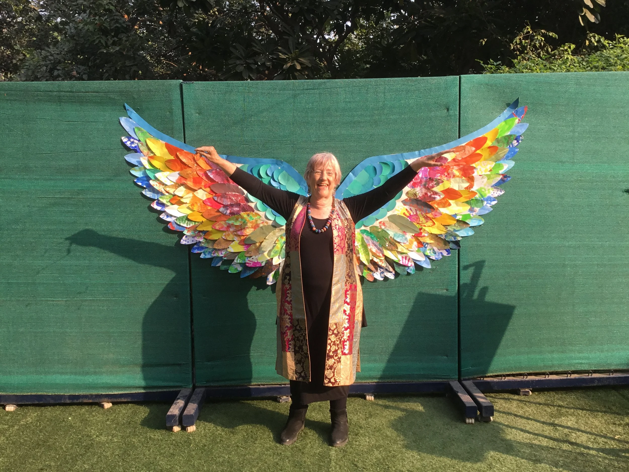 A woman with gray hair, smiling, standing in front of colorful butterfly wings outdoor backdrop, arms outstretched.
