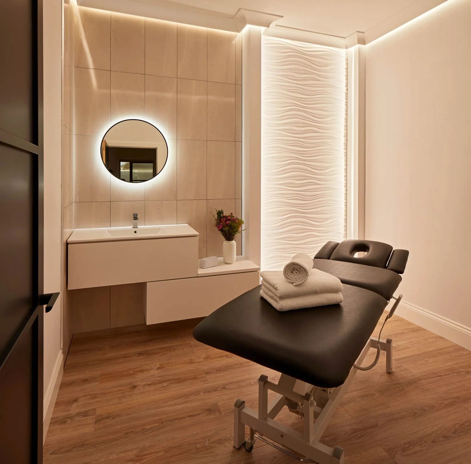 A modern medical or spa room with a black examination chair, rolled white towels, a side table with folded towels, a sink with a mirror, a vase with flowers, and textured wall decor, all illuminated by warm lighting.
