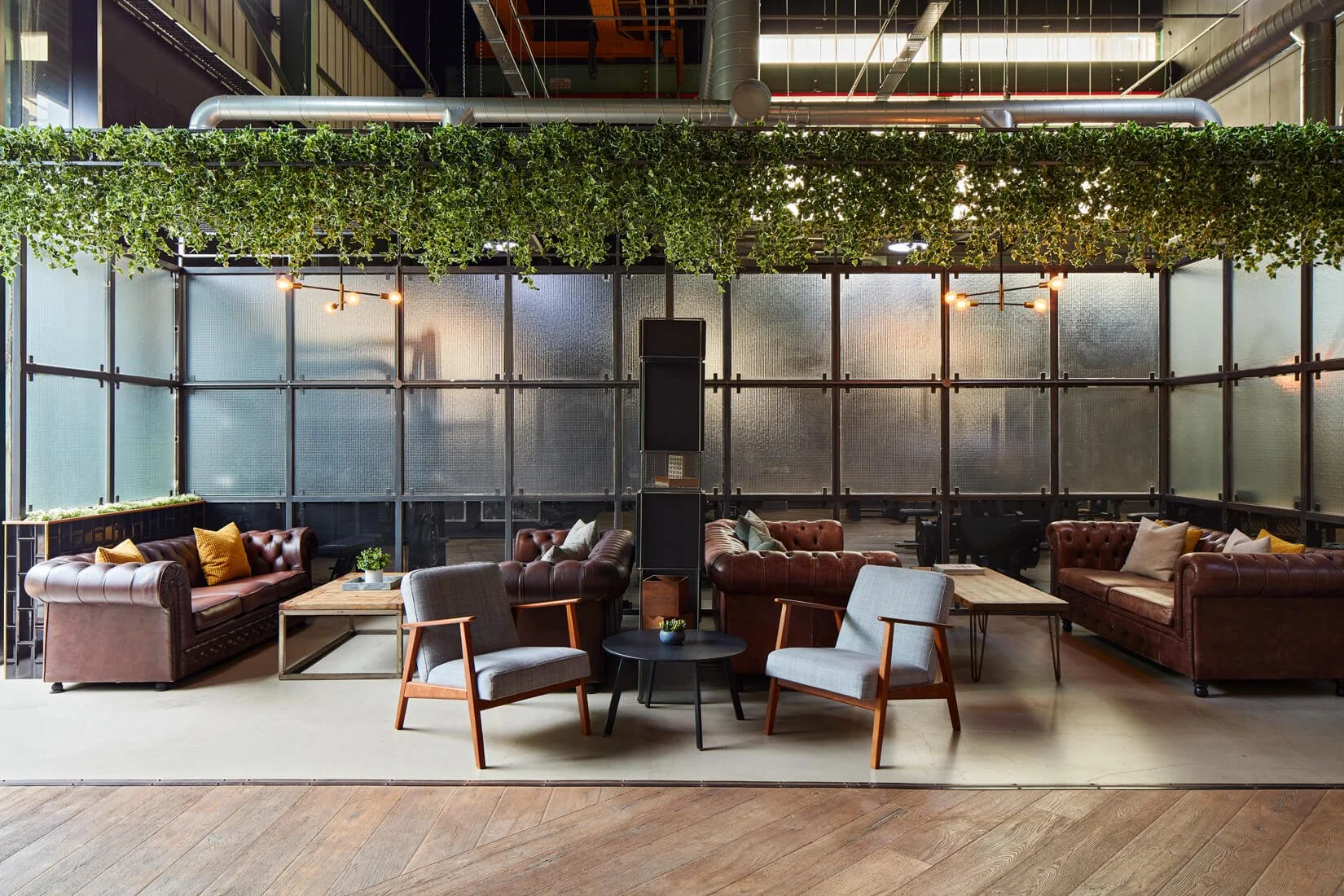 Modern lounge area with leather sofas, armchairs, and small coffee tables, enclosed by a glass partition with frosted panels, and decorated with green hanging plants and contemporary lighting fixtures.