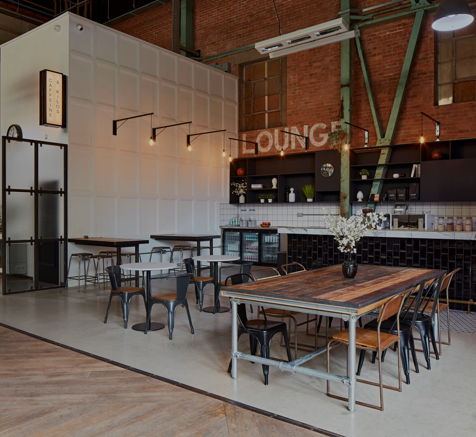 Interior of a modern cafe with a brick wall, exposed green metal beams, black shelving with plants and decor, a white tiled counter, and tables with black chairs, including a large wooden table with a flower vase.