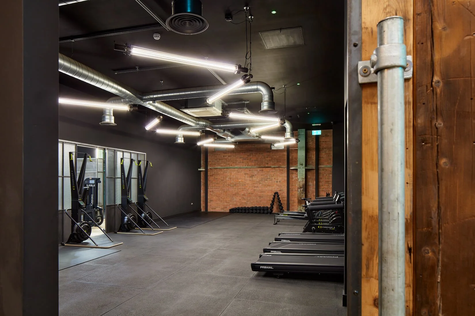 Modern gym with black walls, exposed brick, and ceiling piping, equipped with Concept2 rowing machines, treadmills, and weights.