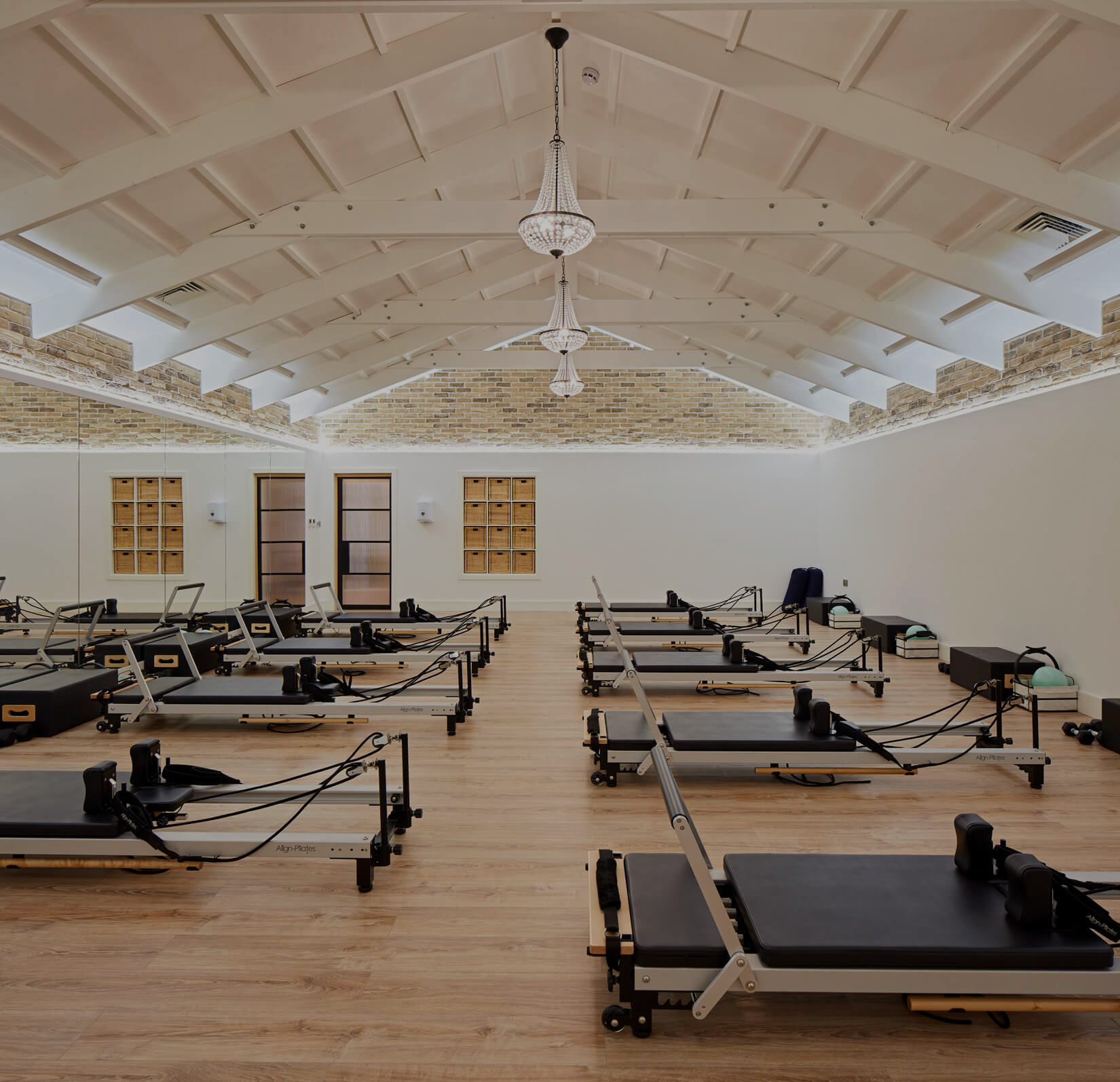 Pilates studio with multiple Reformers arranged on a wooden floor, brick walls, white ceiling with chandeliers, large mirror, and windows.