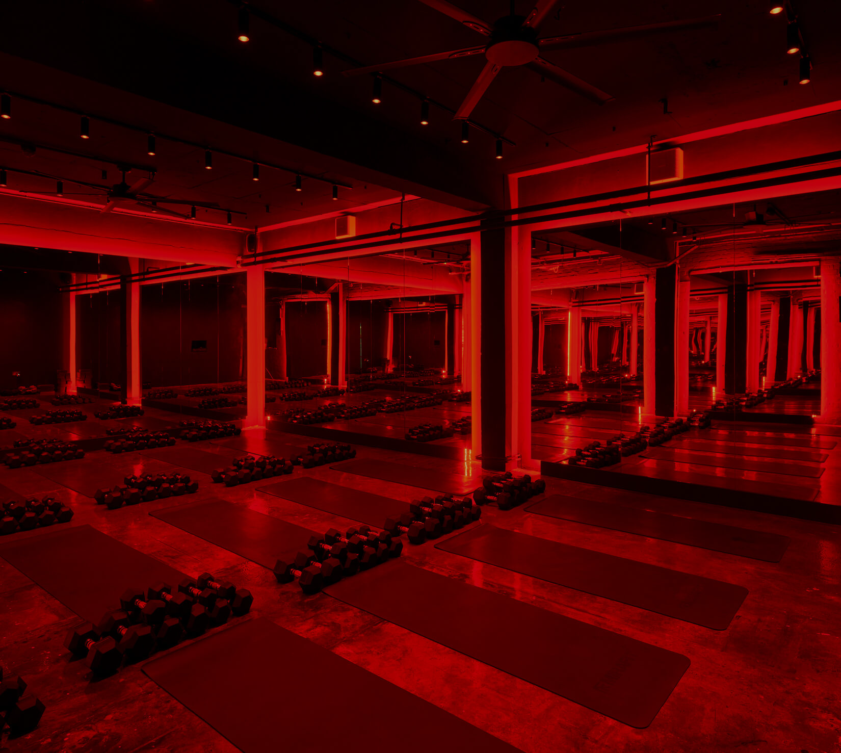 A dark gym room with red lighting, mirrors on the walls, and workout mats with dumbbells on top of the mats.