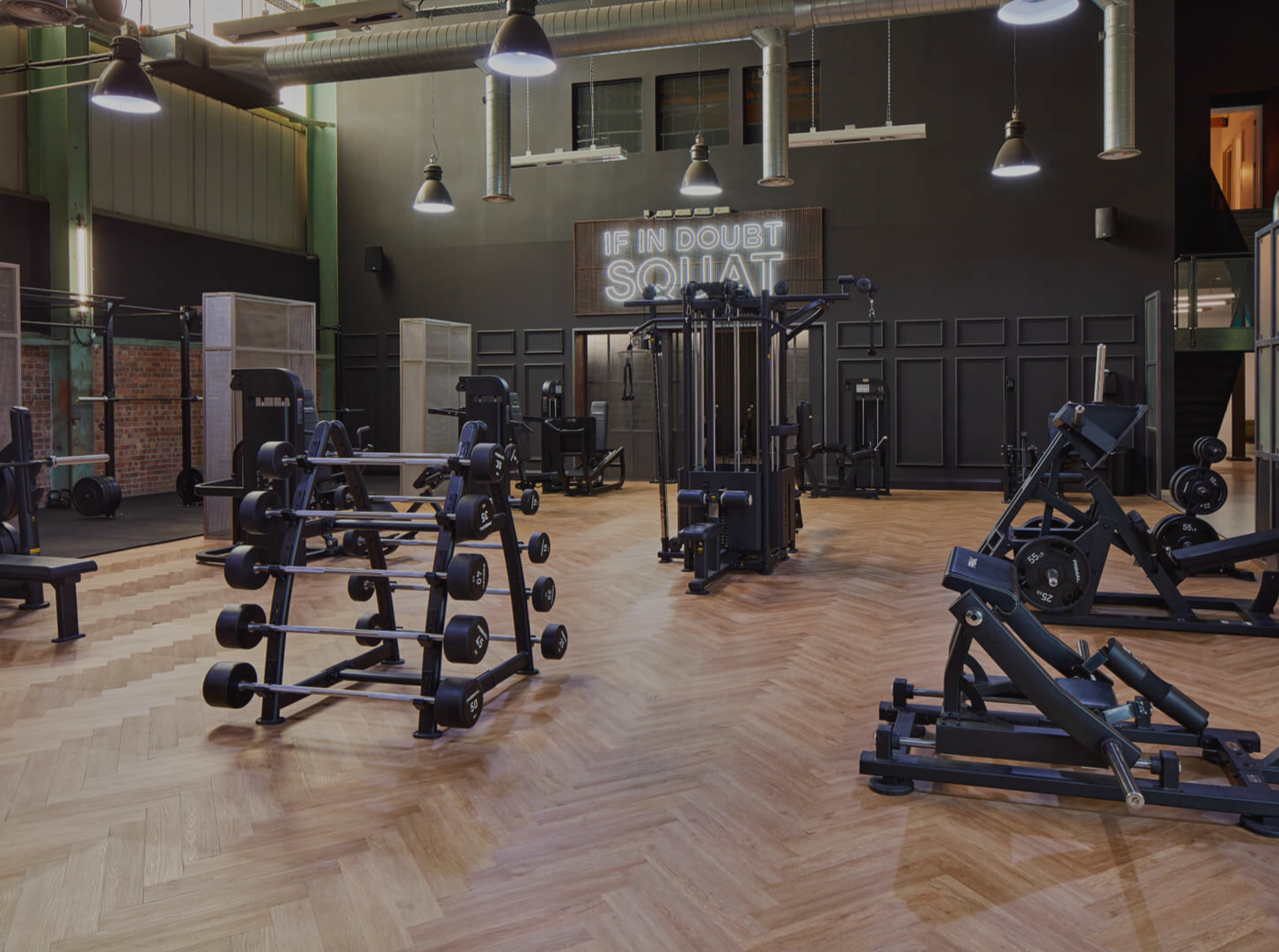 Modern gym with various workout equipment including dumbbells, weight machines, and cardio machines; black and wooden decor with a neon sign 'IN DOUBT SQUAT' on the wall.