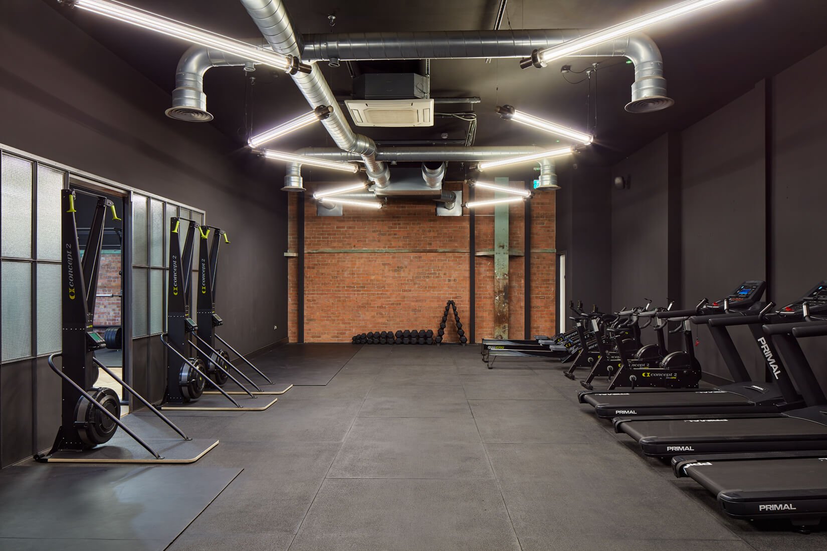 Empty gym with black walls, exposed ductwork ceiling, and brick wall in the background. Equipment includes rowing machines, treadmills, and weight racks, all neatly arranged.