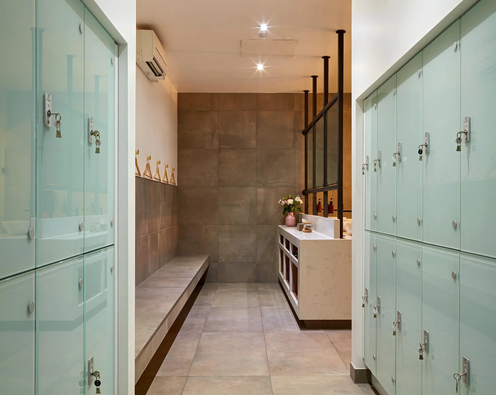 Locker room with mint-colored lockers, a bench, a wall-mounted air conditioning unit, a small counter with a pink vase and flowers, and modern lighting.