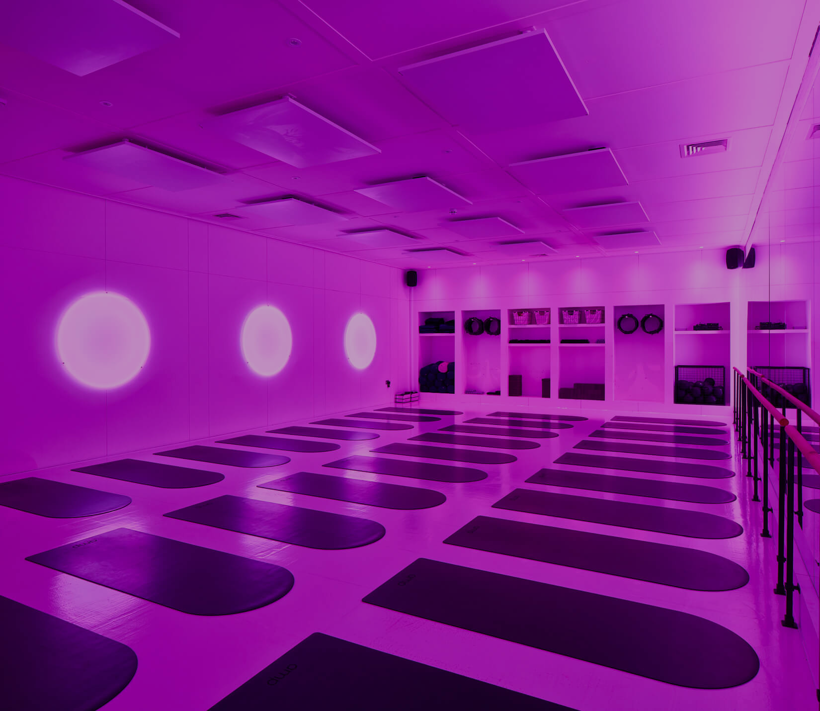 Yoga class studio with purple lighting, yoga mats on the floor, and shelves with fitness equipment.