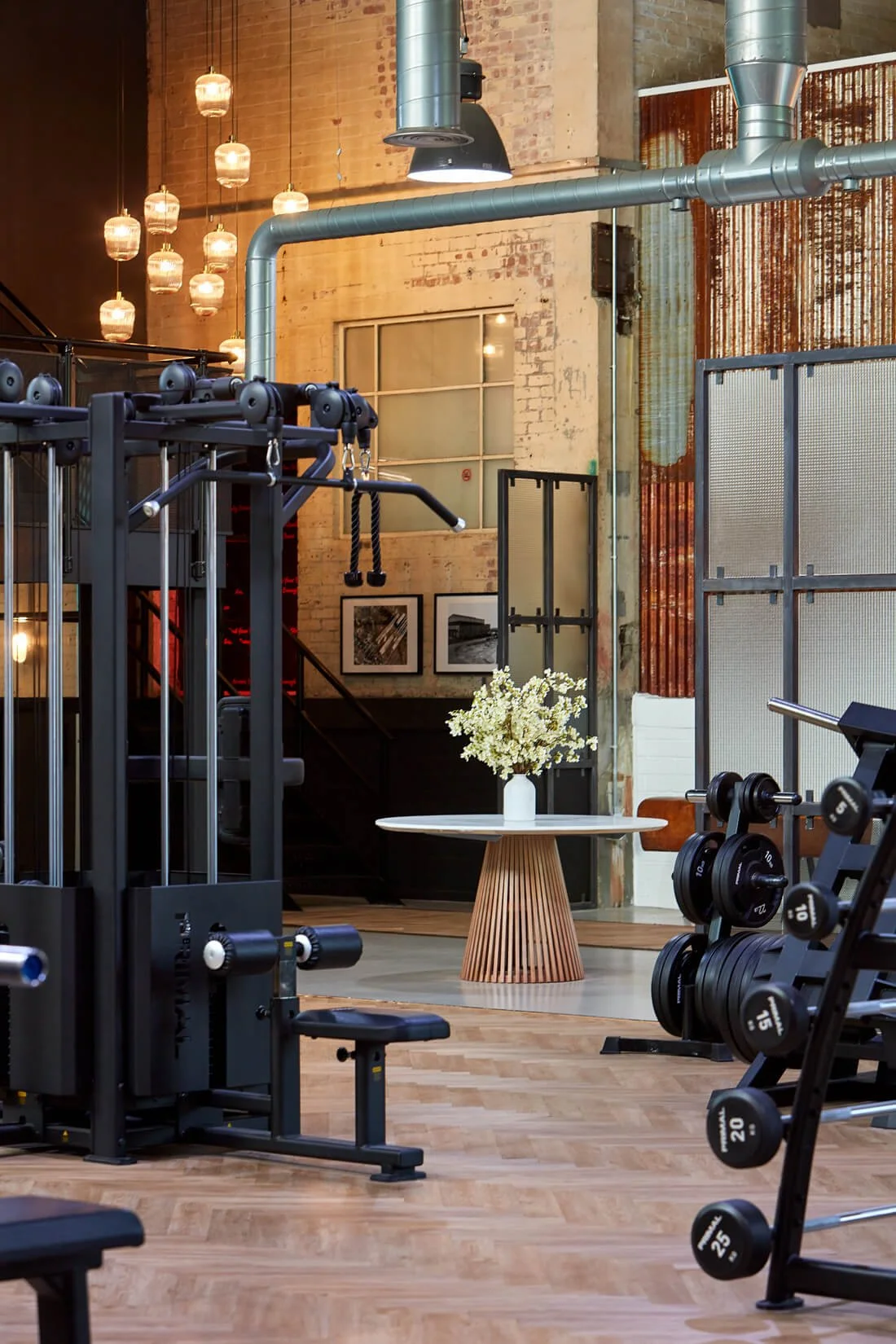 Modern gym with weightlifting equipment, a white flower arrangement on a wooden table, and industrial decor including exposed brick and metal piping.