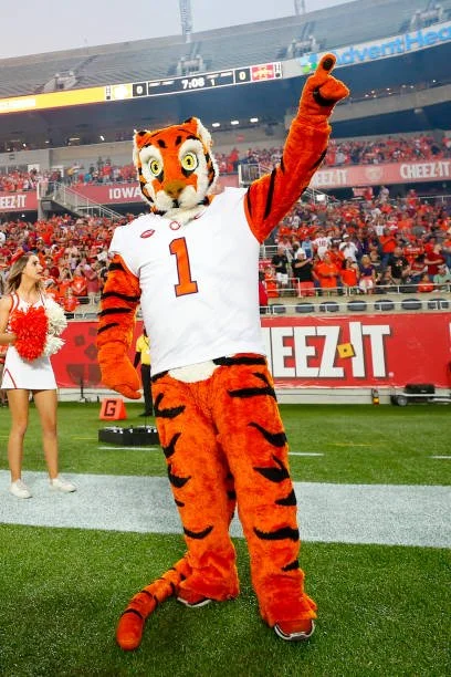 Tiger mascot wearing a white football jersey with the number 1, standing on a football field, waving with one arm raised, during a game in a stadium filled with spectators.