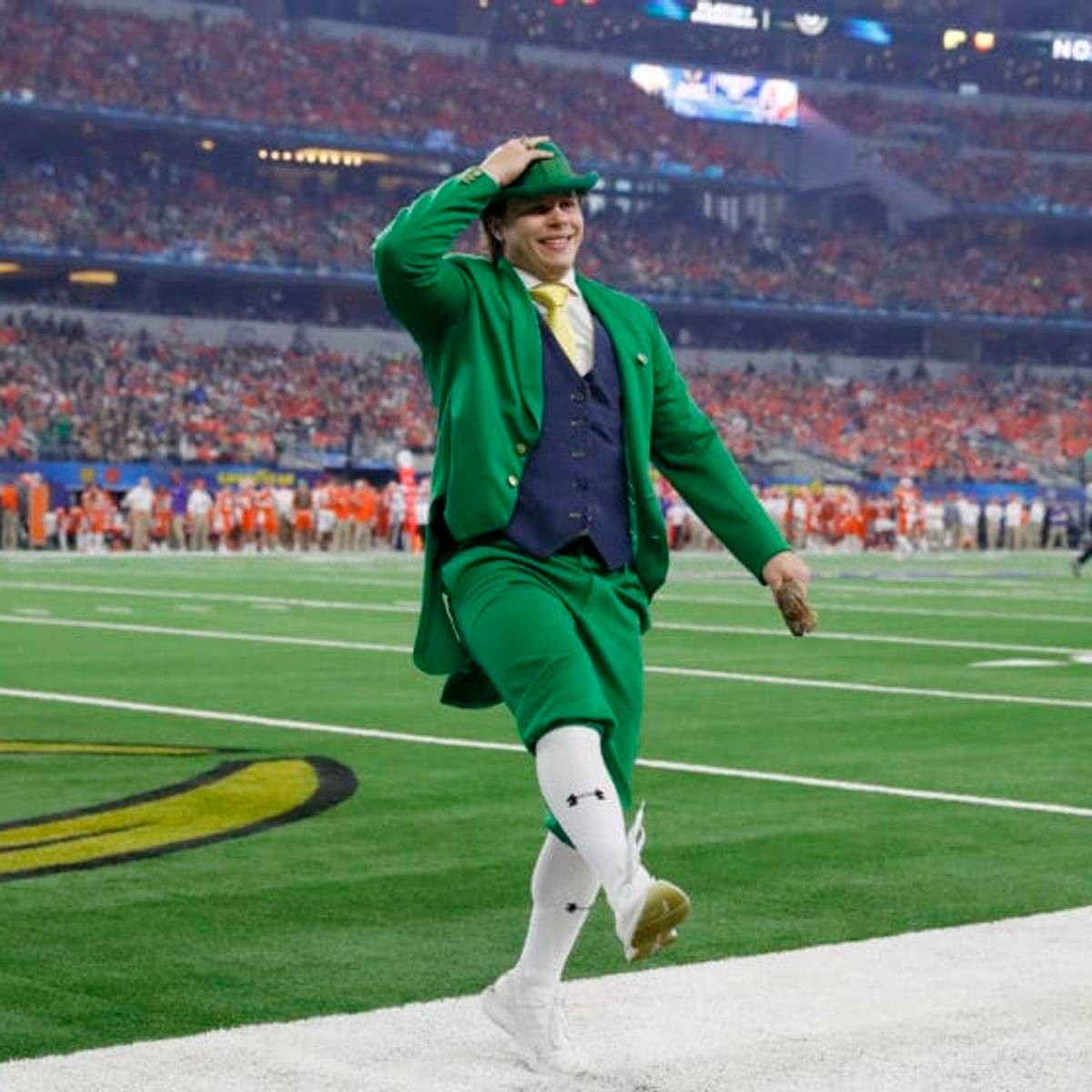 A person dressed as a leprechaun in a bright green suit, black vest, and white knee-high socks, smiling and running across a football field in a stadium filled with spectators.