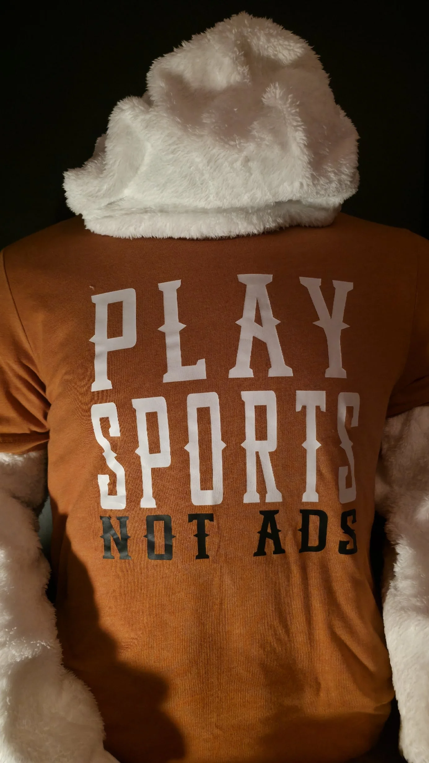 Person wearing a white fluffy hat, a brown shirt with white and black text that reads 'PLAY SPORTS NOT AD'S', and a plush white furry coat.