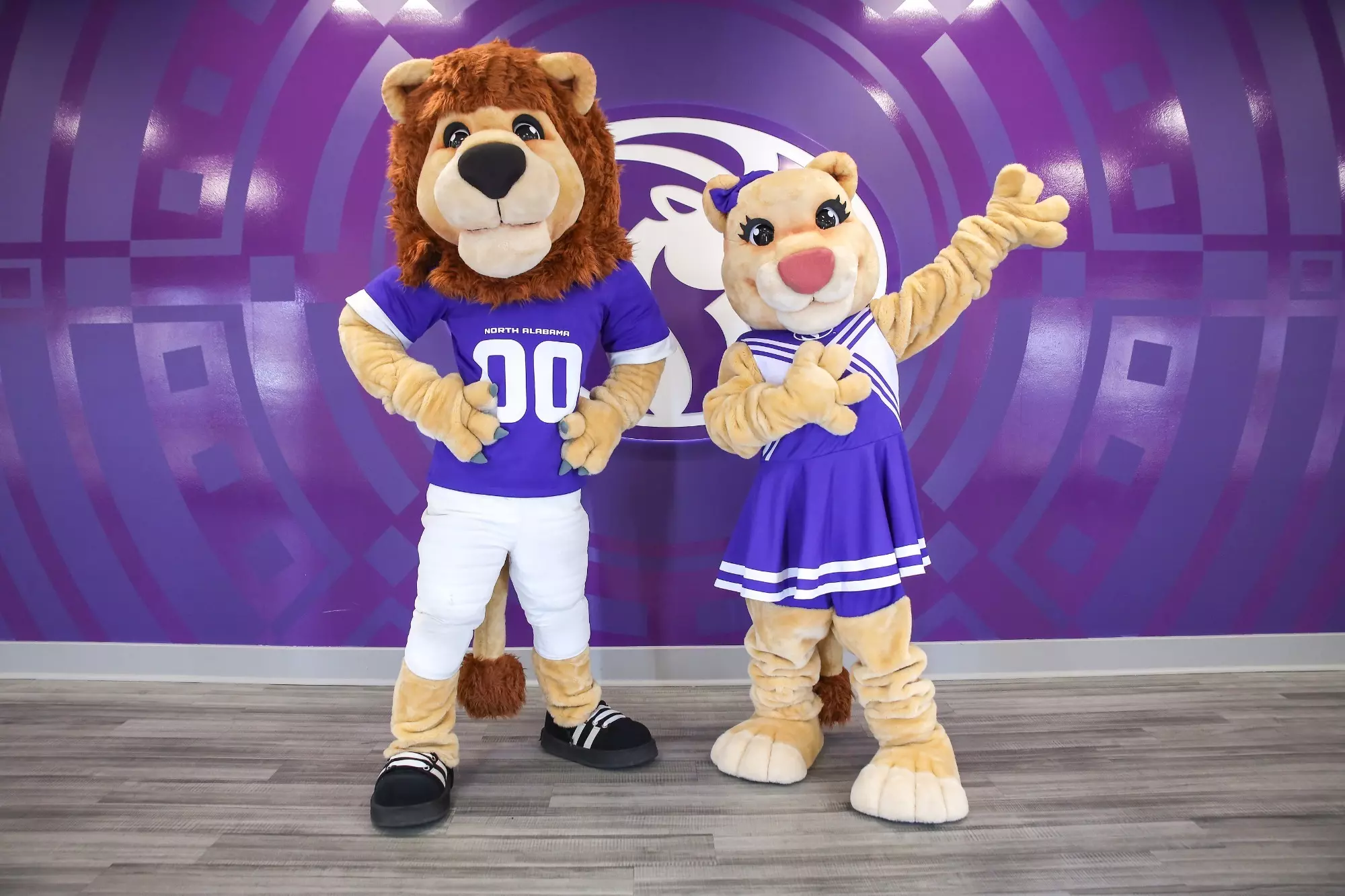 Two people dressed in lion costumes posing in front of a purple background with a white lion logo. The person on the left wears a blue jersey, white pants, and black sneakers. The person on the right wears a blue and white striped dress and a blue bow on their head.