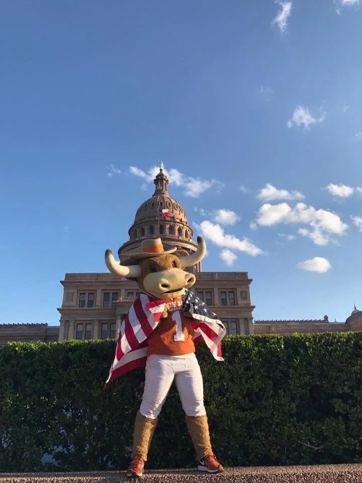 Costumed character of a friendly bull wearing a cowboy hat, American flag cape, orange shirt, white pants, and boots, standing in front of the Texas State Capitol building under a blue sky with clouds.
