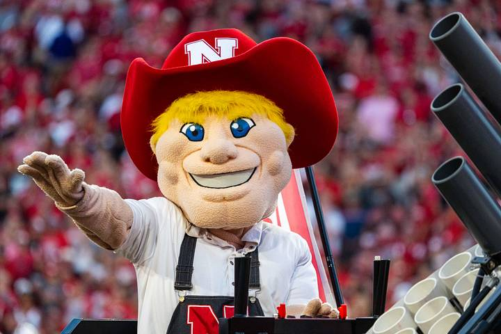 A mascot with blonde hair, a big smile, and big blue eyes, wearing a red hat with a white N, and a white shirt, standing in front of a crowd at a sports stadium.