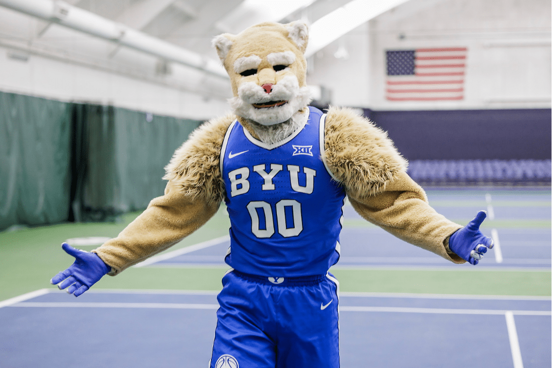 College mascot in a lion costume standing on a tennis court with arms outstretched and wearing BYU sports uniform.